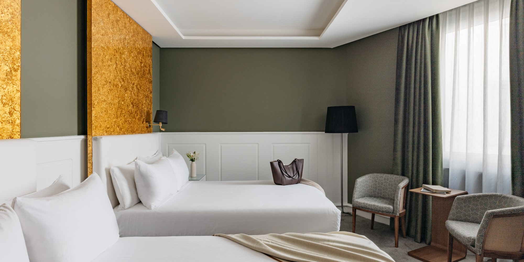 Suite hotelera con dos camas, decoración en verde oliva y detalles dorados.