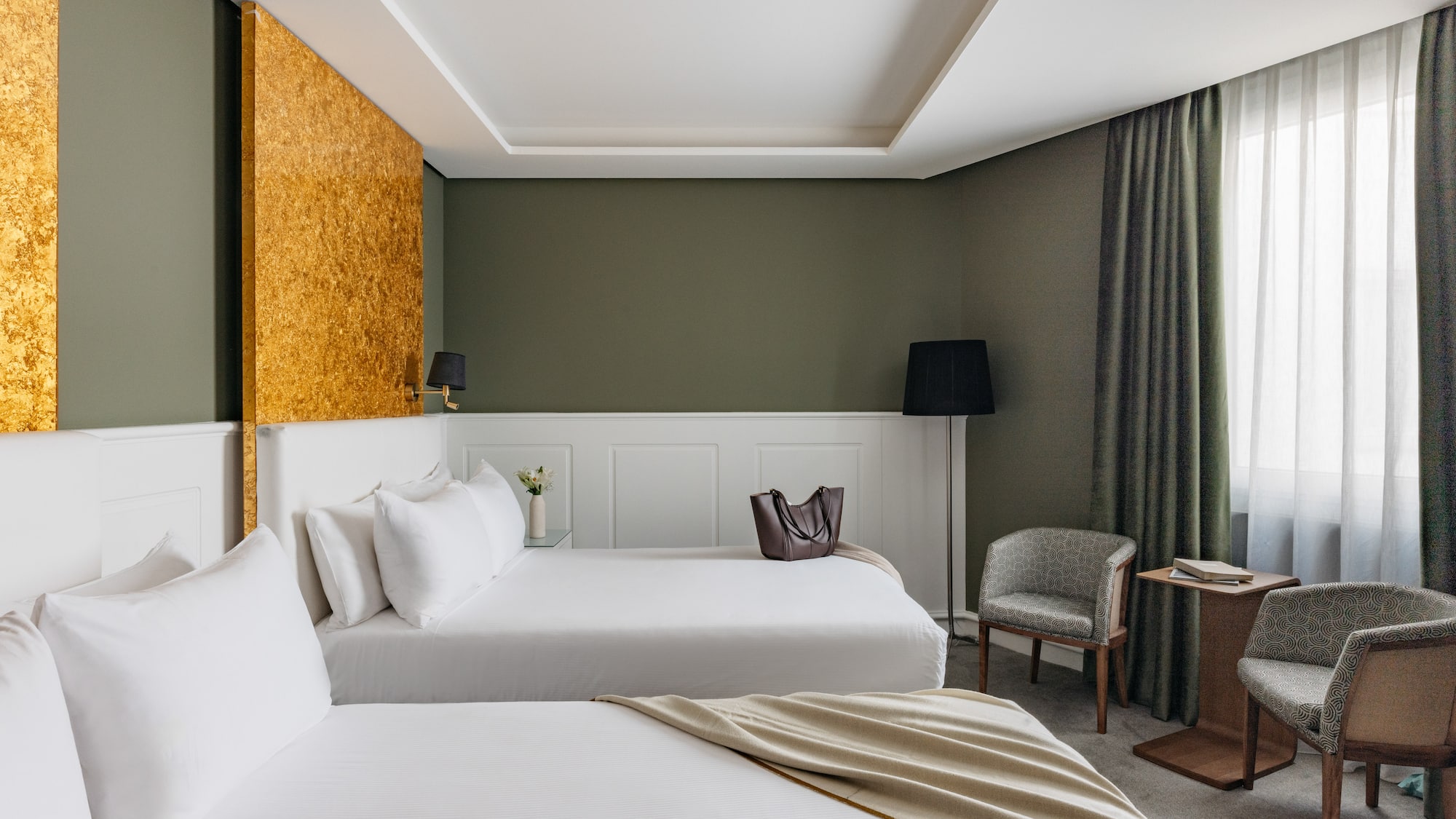 Suite hotelera con dos camas, decoración en verde oliva y detalles dorados.