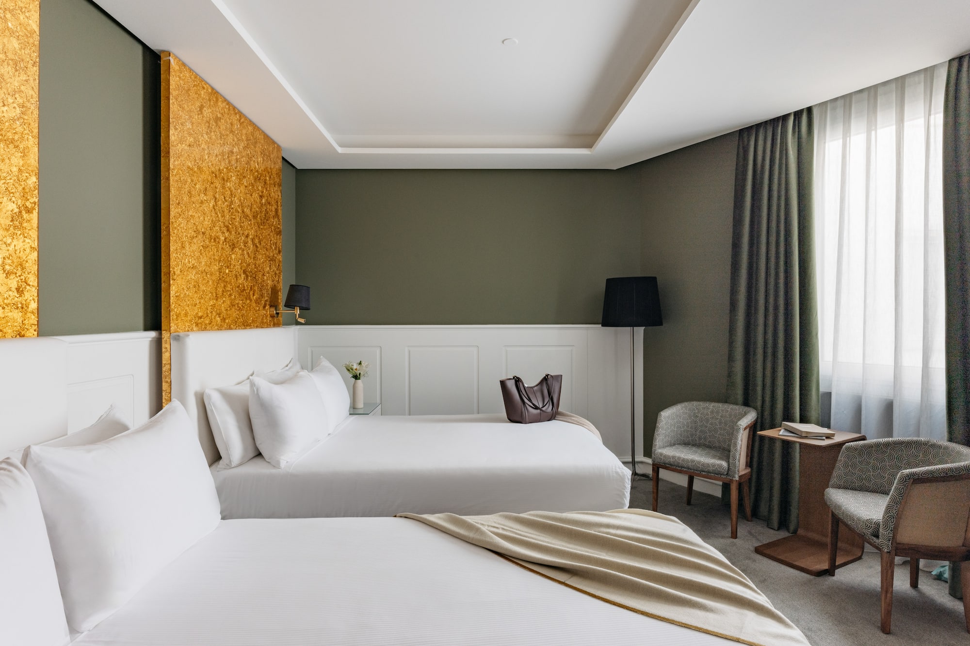 Suite hotelera con dos camas, decoración en verde oliva y detalles dorados.