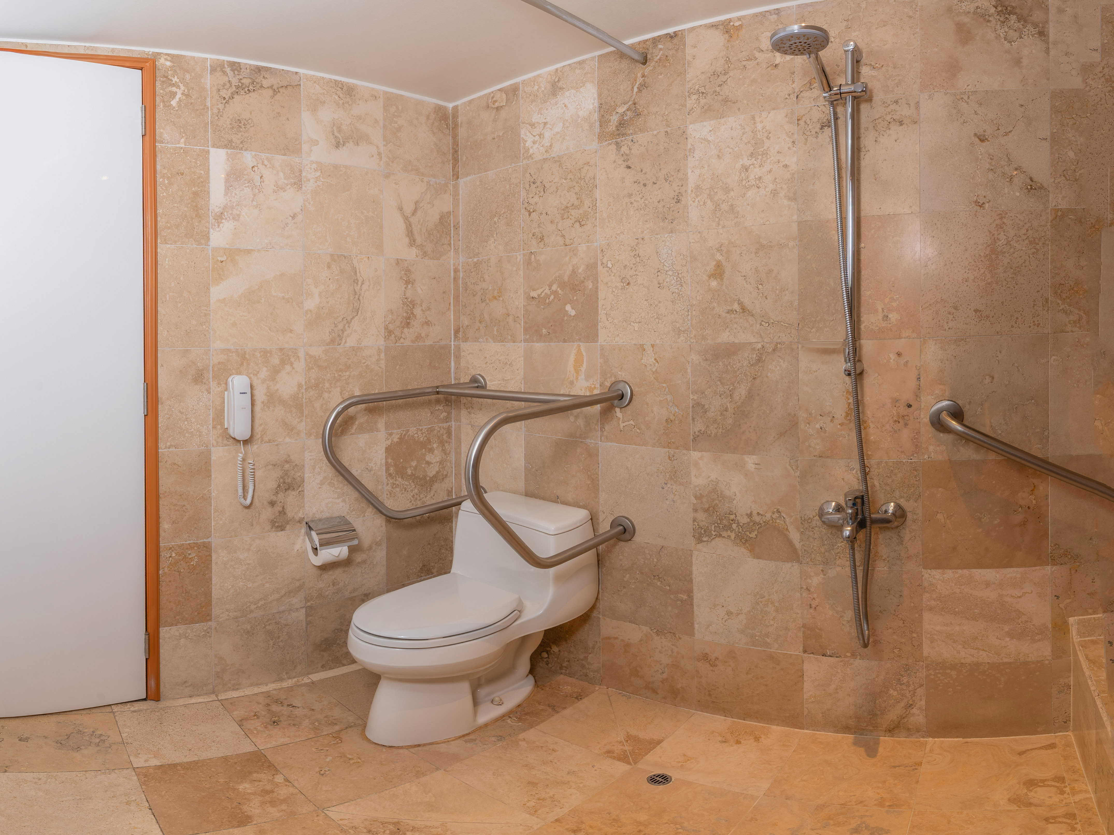 Baño accesible con inodoro, barras de apoyo, ducha y azulejos beige.