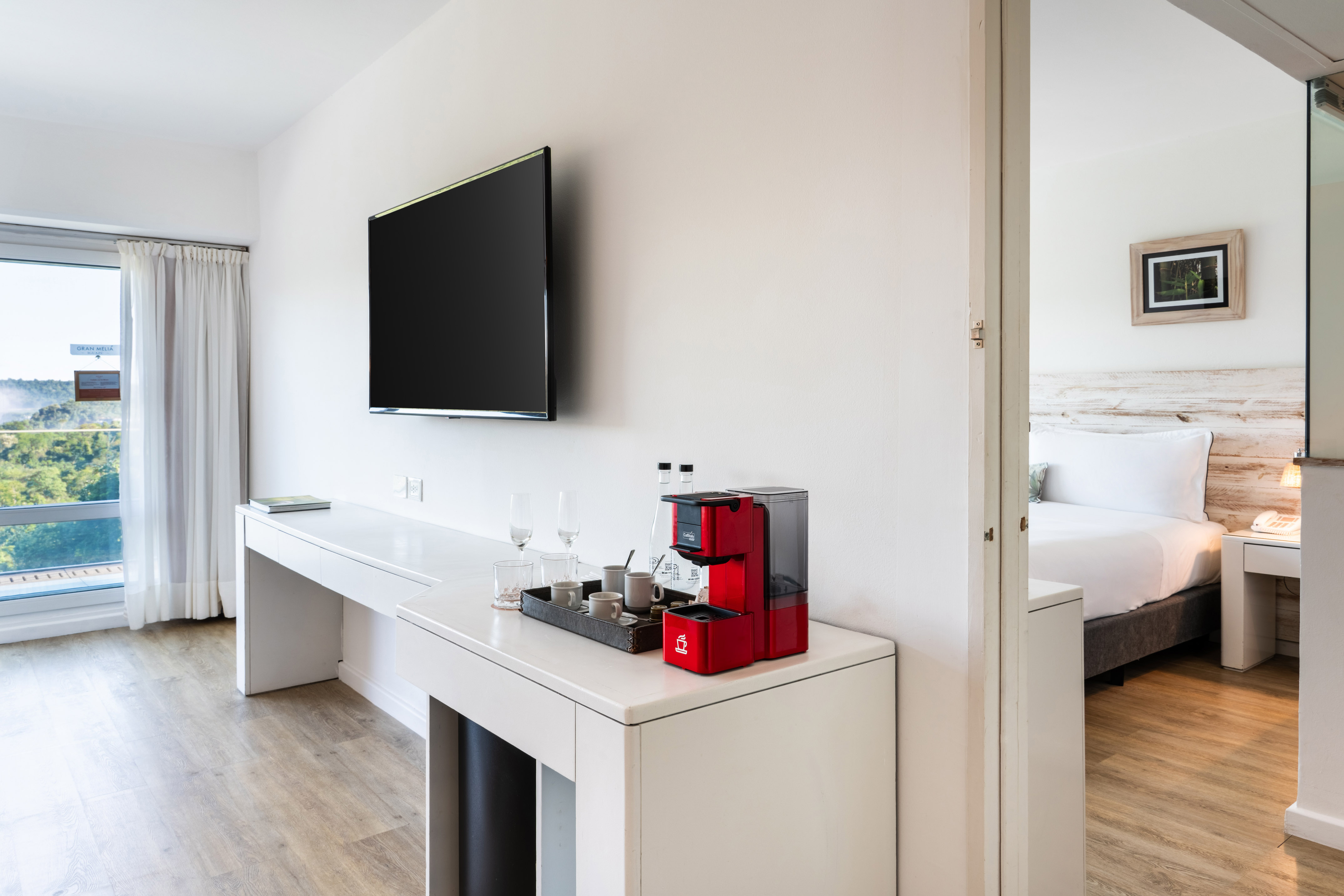 Modern hotel suite: desk, wall TV, coffee station, scenic view, and adjoining bedroom.