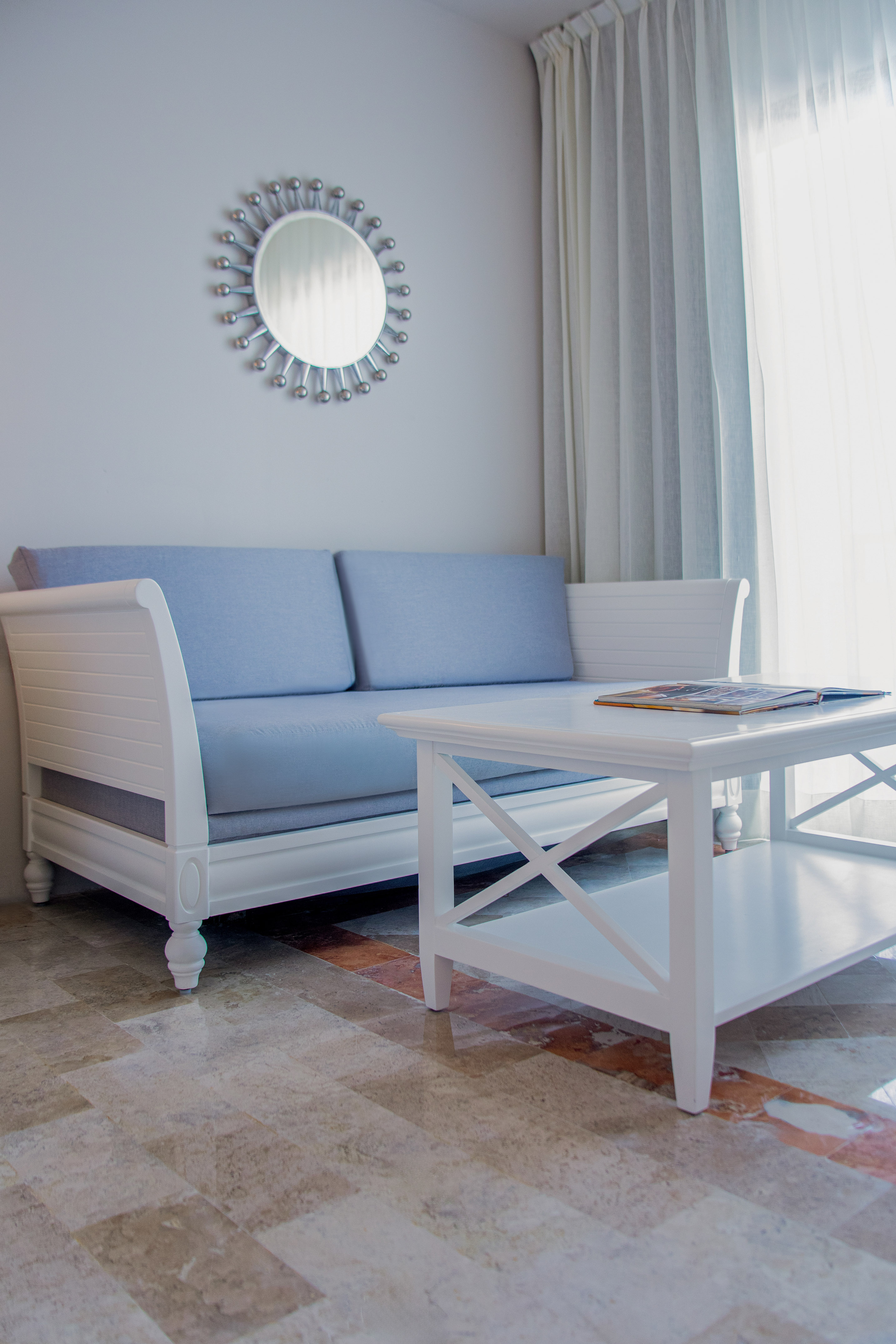 White sofa, blue cushions, coffee table, sunburst mirror in bright room.