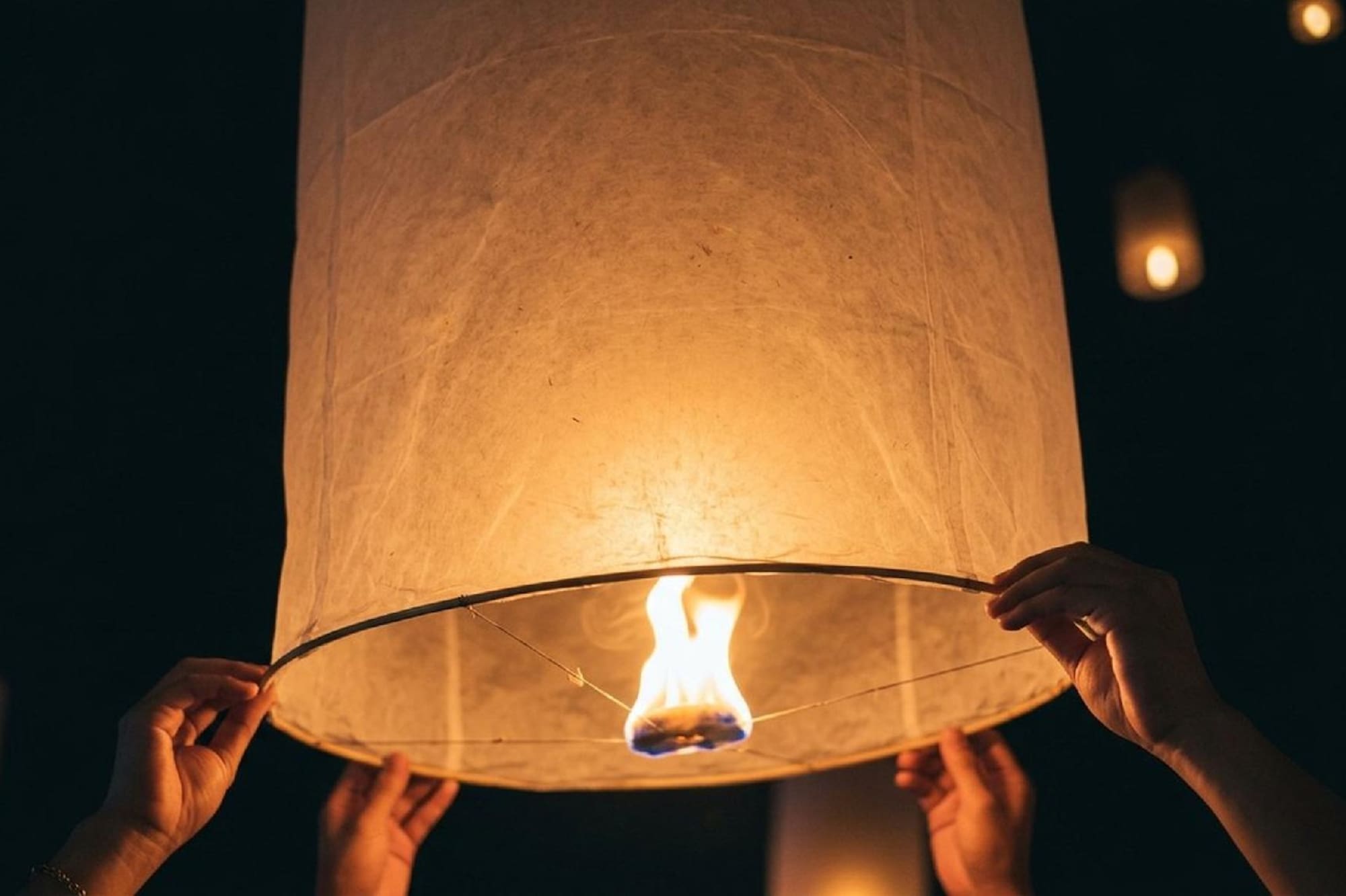 Hands holding a lit paper sky lantern with a flame at night.