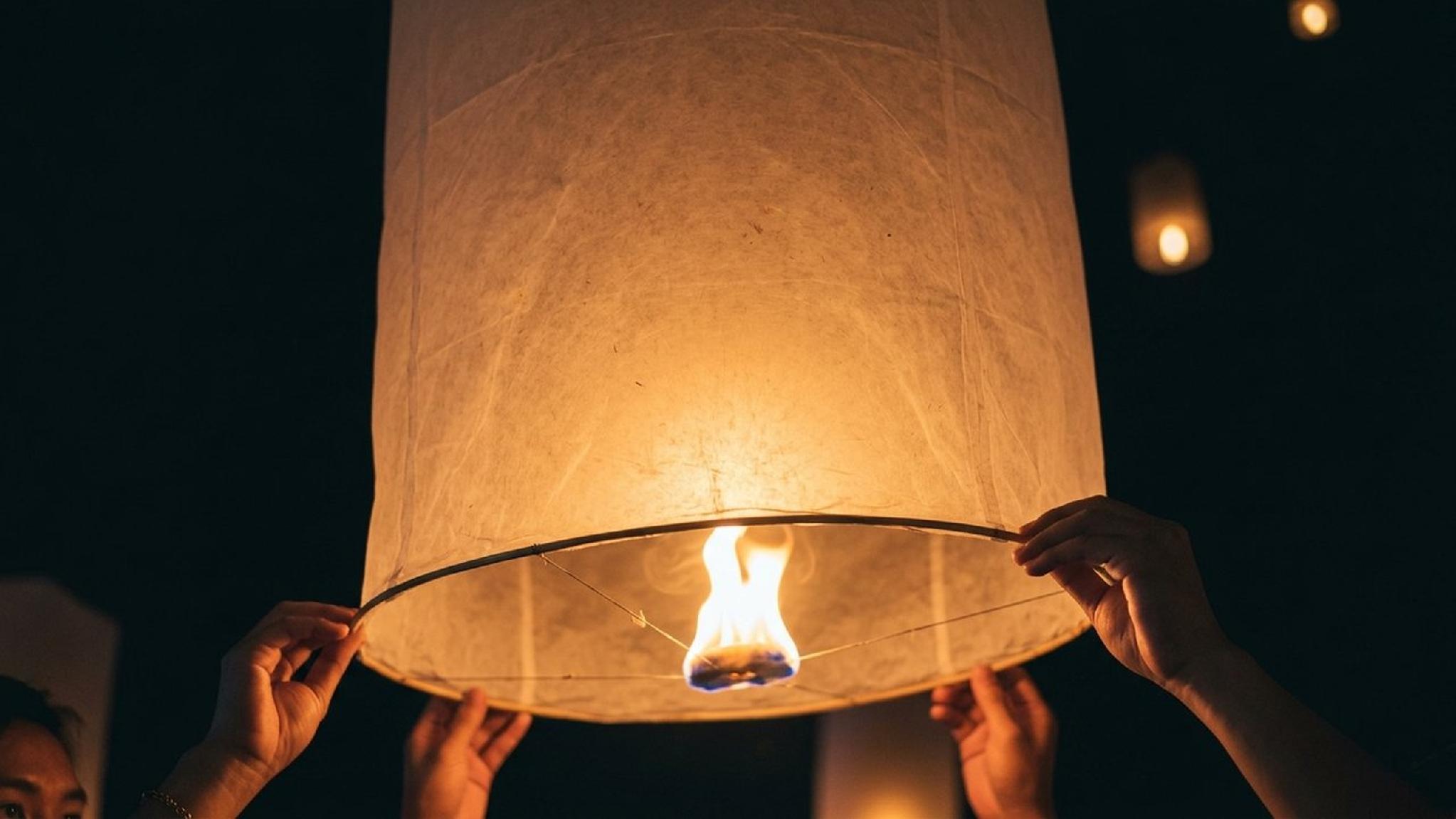 Hands holding a lit paper sky lantern with a flame at night.