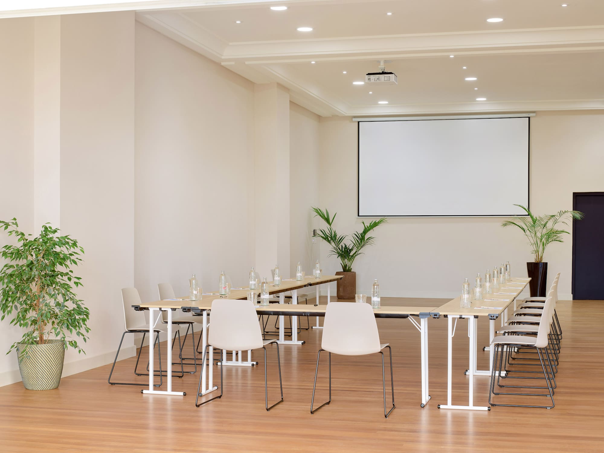 Bright meeting room with U-shaped table, projector screen, and potted plants.
