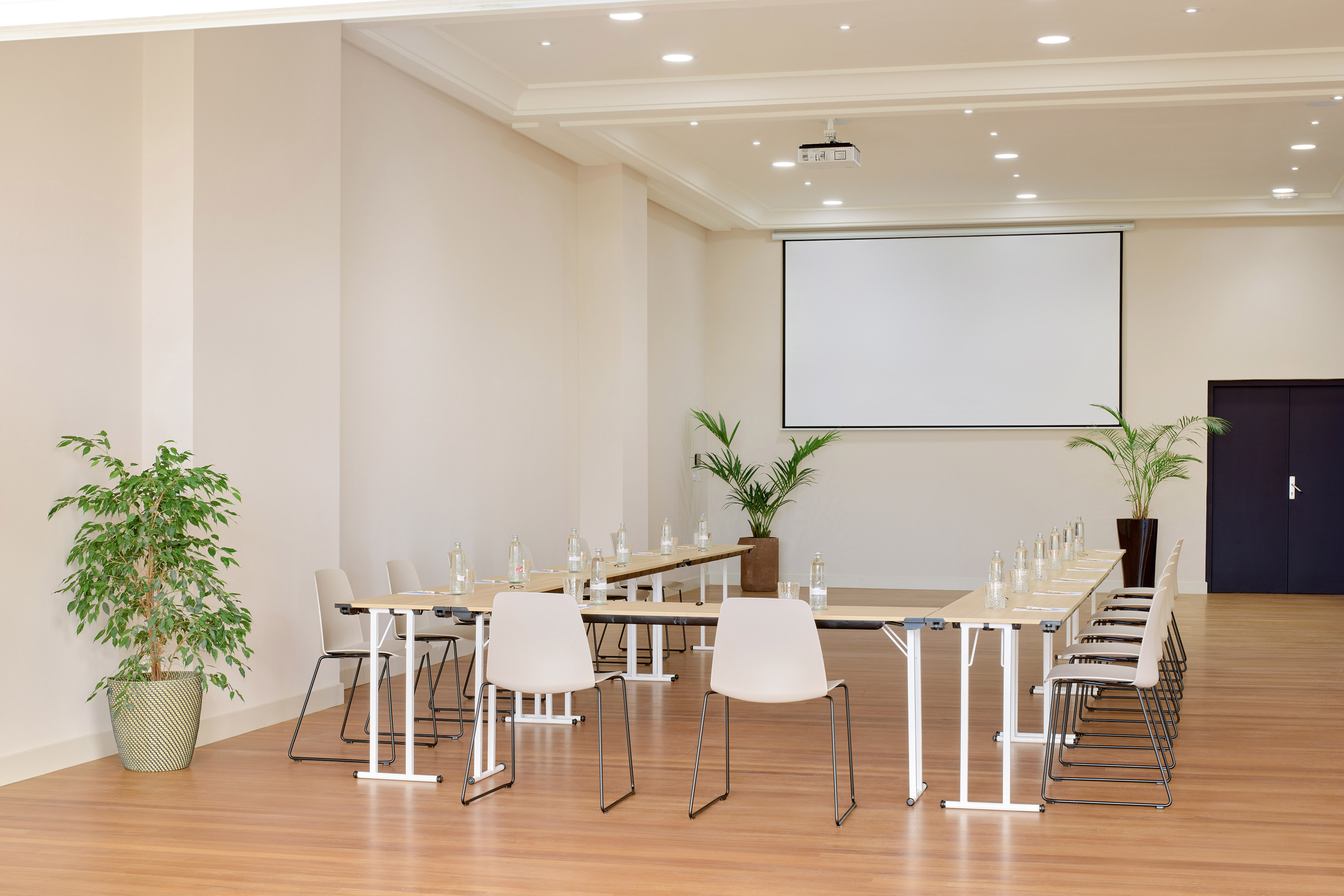 Bright meeting room with U-shaped table, projector screen, and potted plants.
