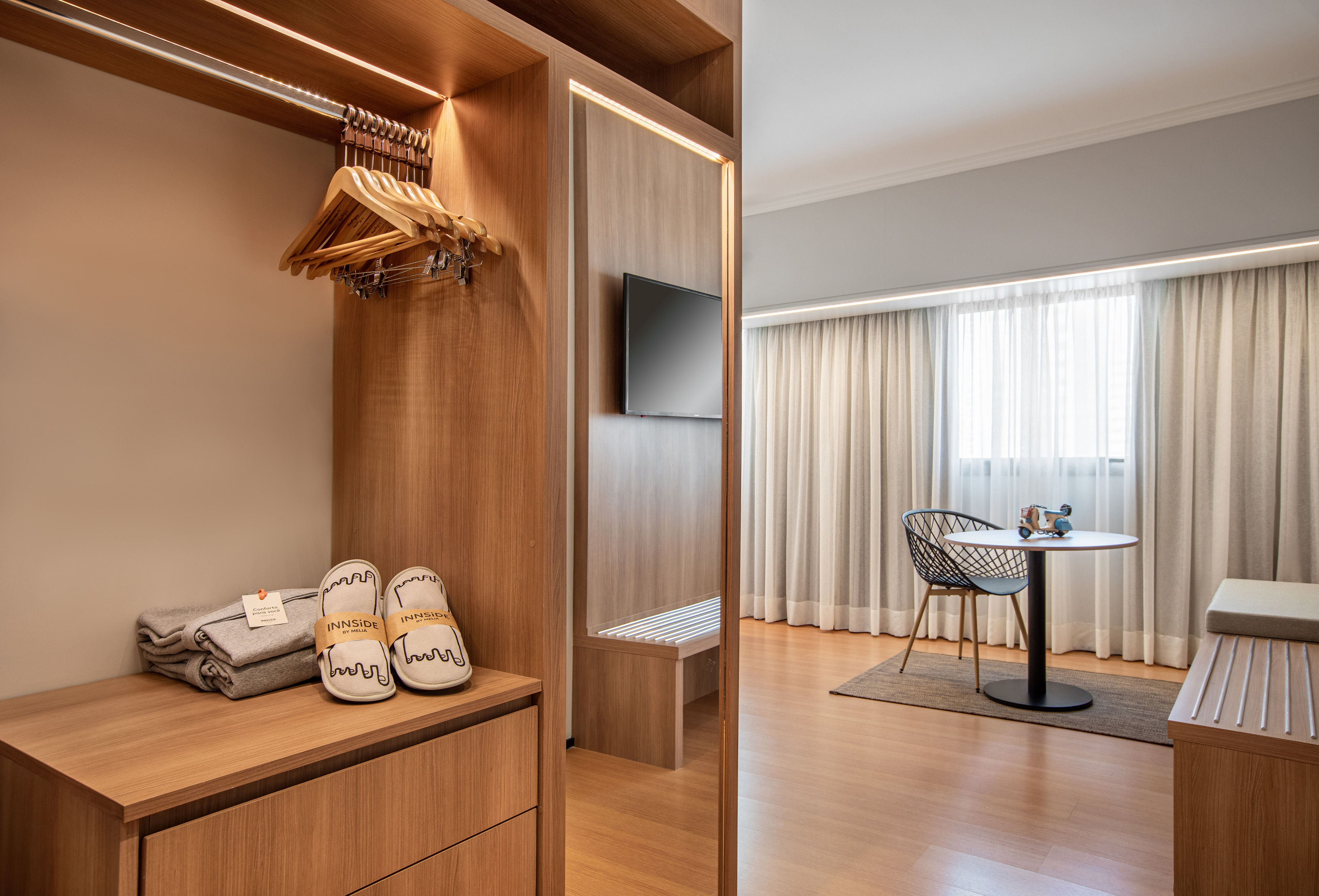 Modern hotel room with illuminated wooden closet, slippers, TV, and window.