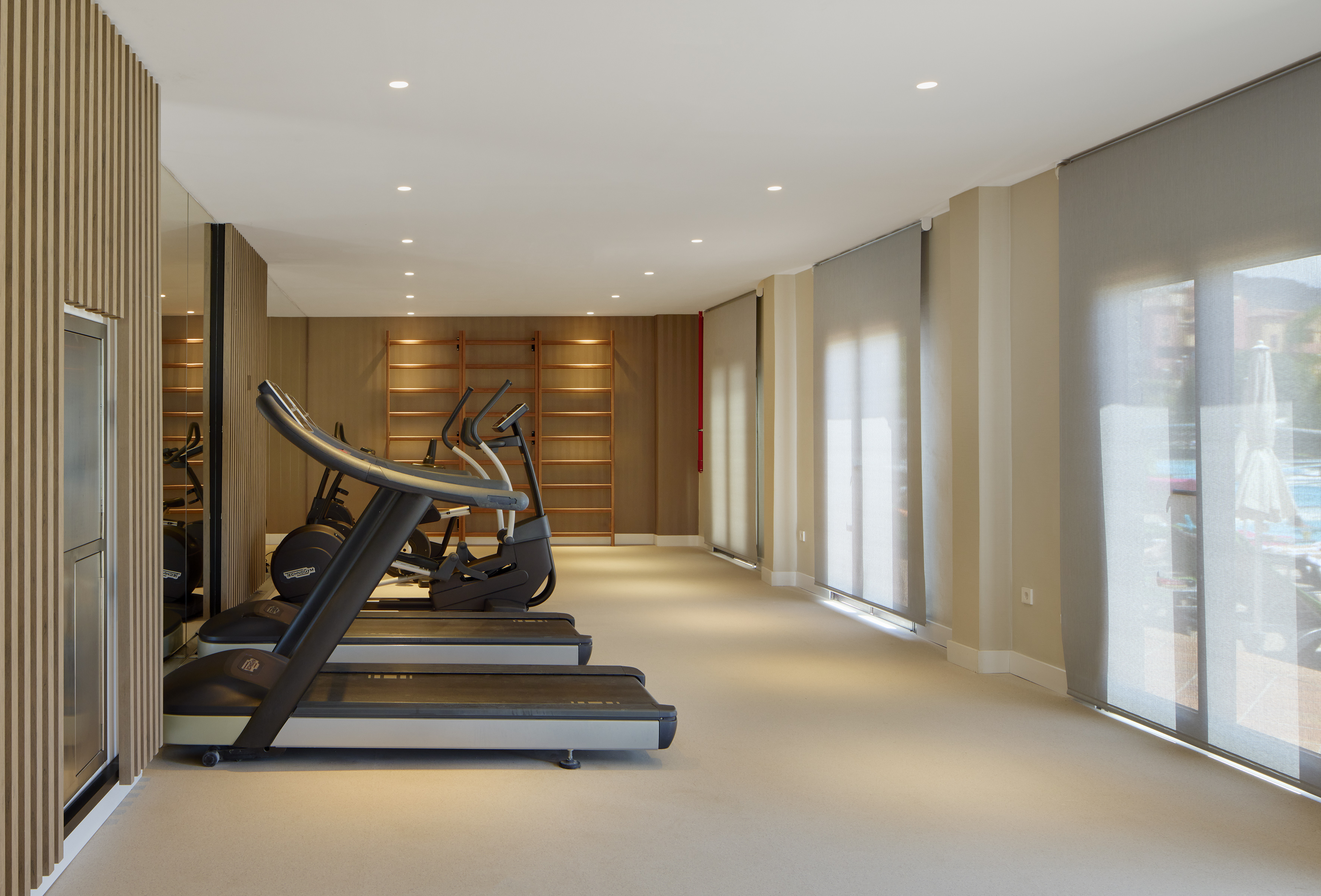 Modern hotel gym with treadmills, ellipticals, wood walls, and pool view.