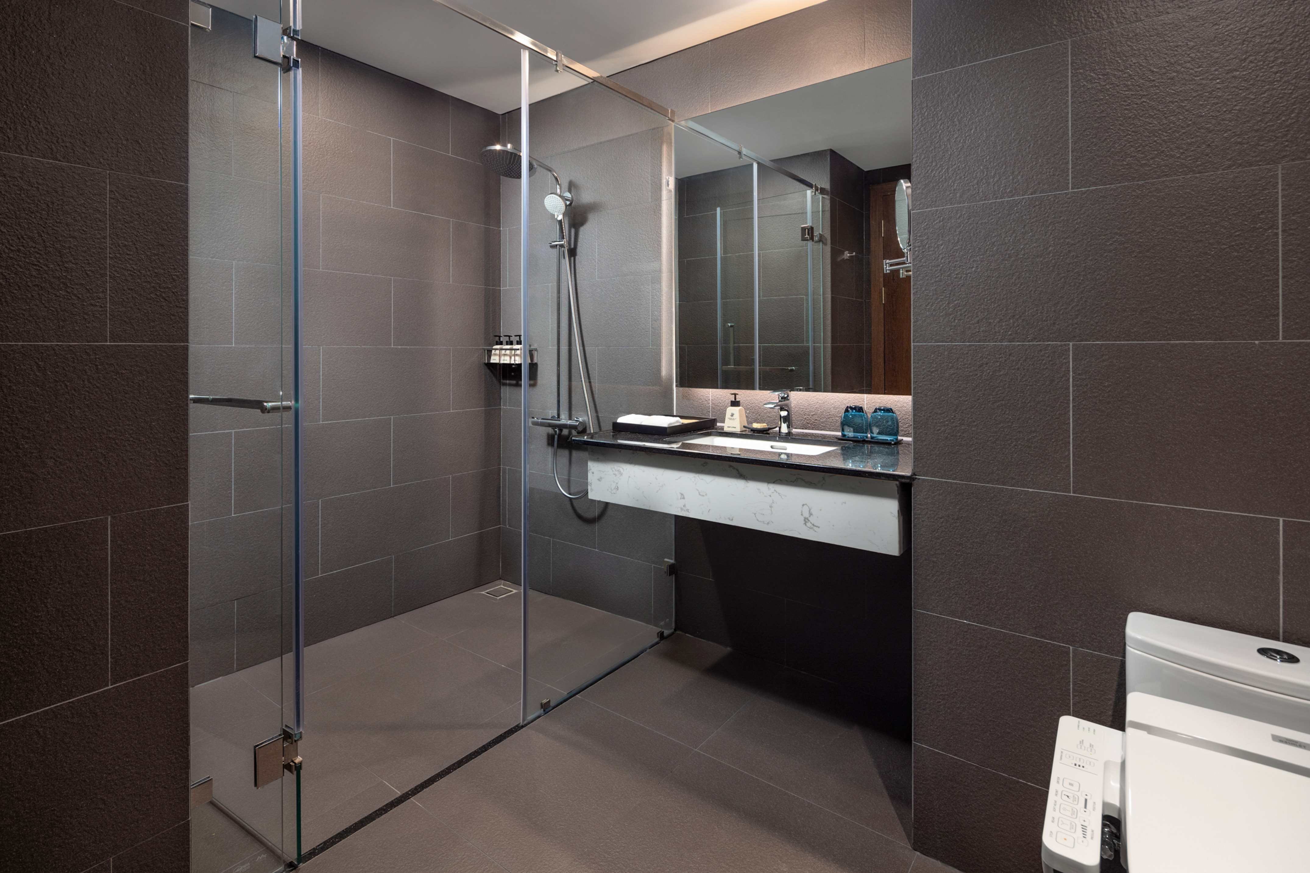 Modern hotel bathroom with dark grey tiles, glass shower, and vanity.