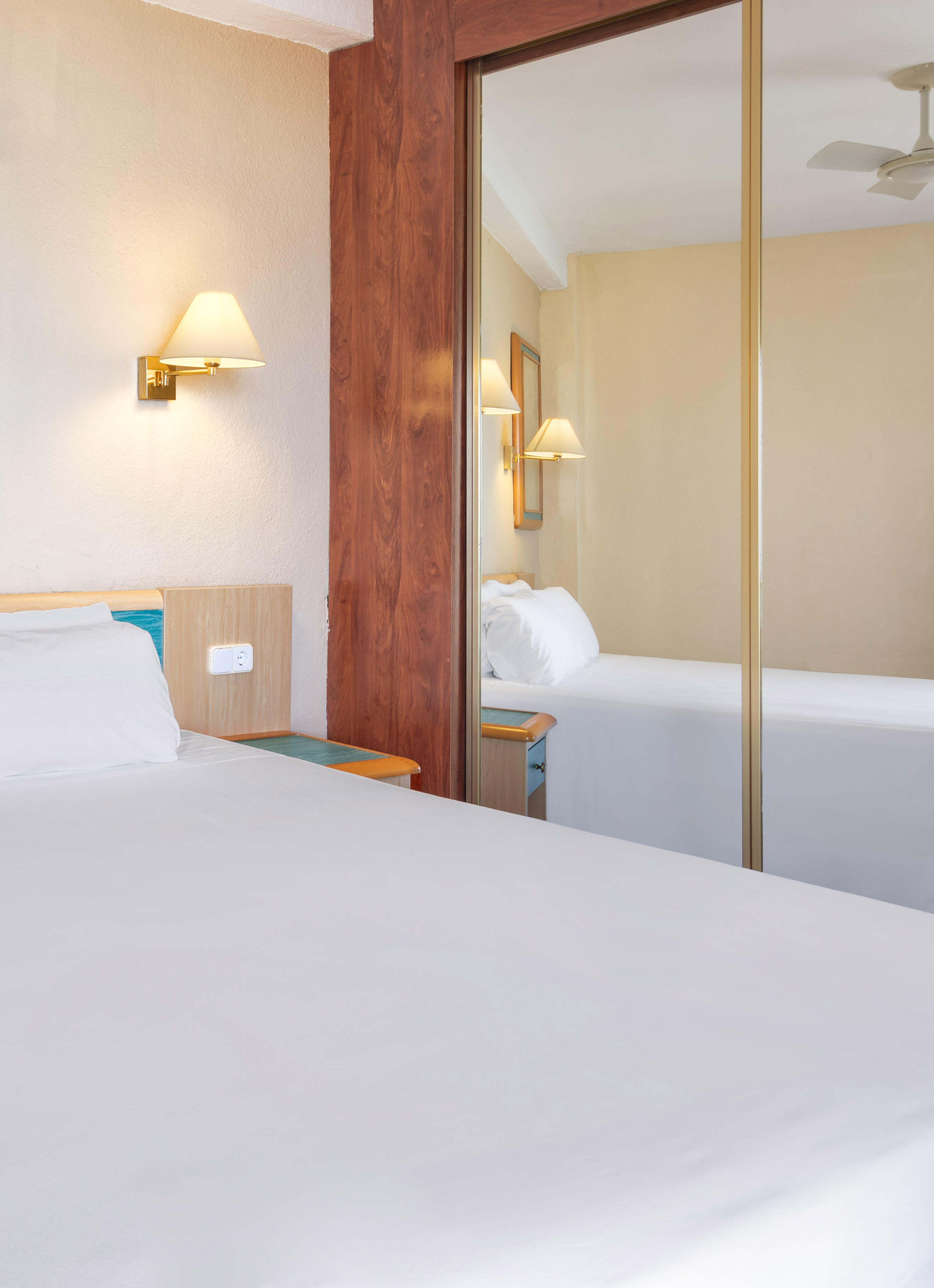 Hotel bedroom with white bed, golden lamp, and mirrored closet reflecting the room.