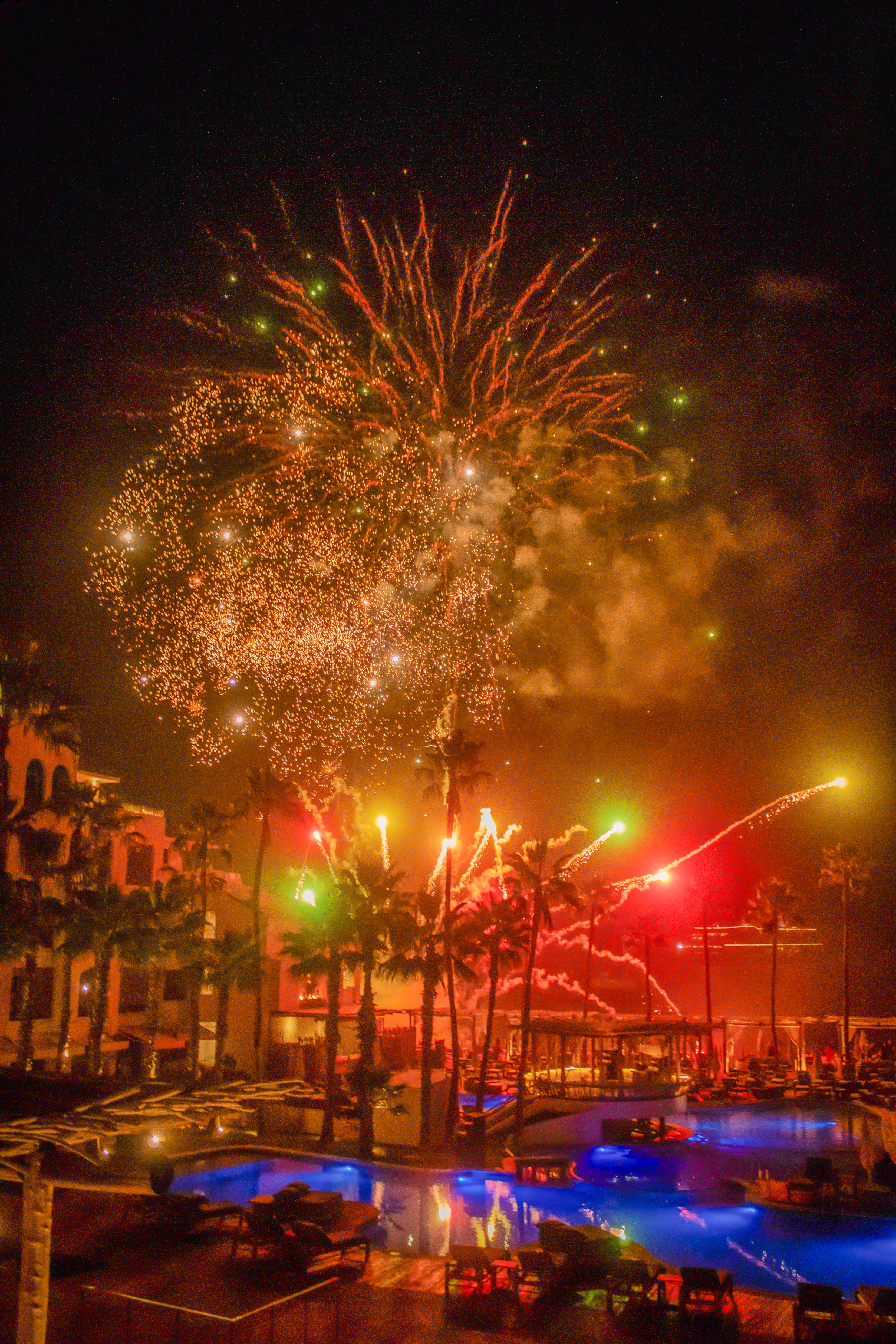 Colorful fireworks over a resort pool and palm trees at night.