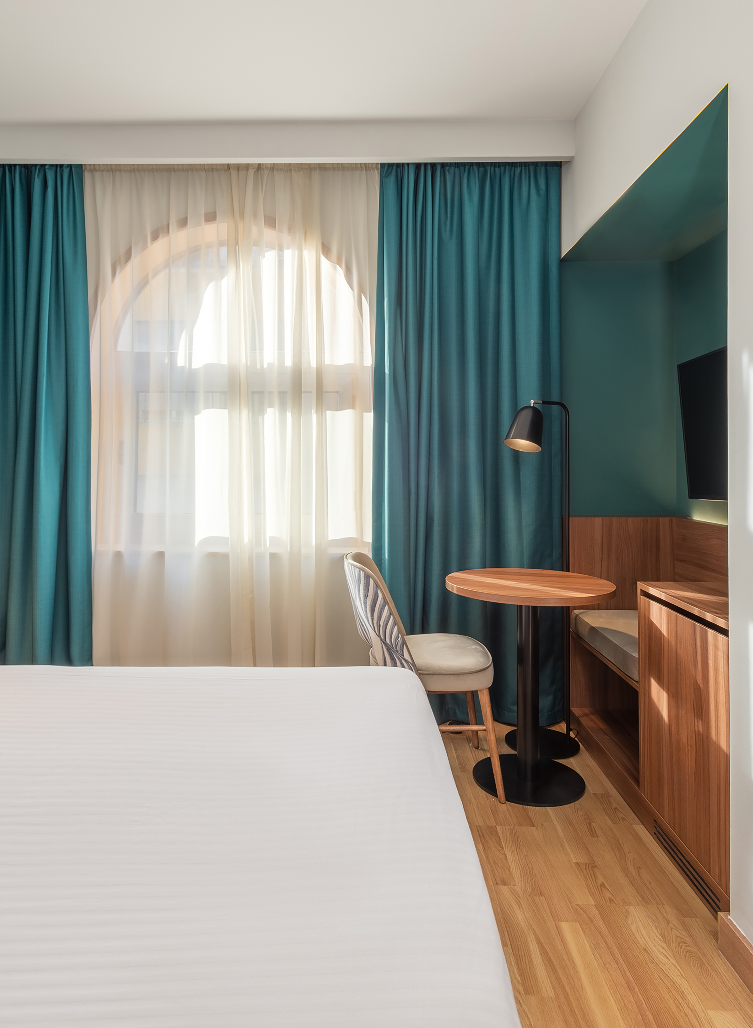 Modern hotel room with white bed, arched window, teal curtains, wooden table.