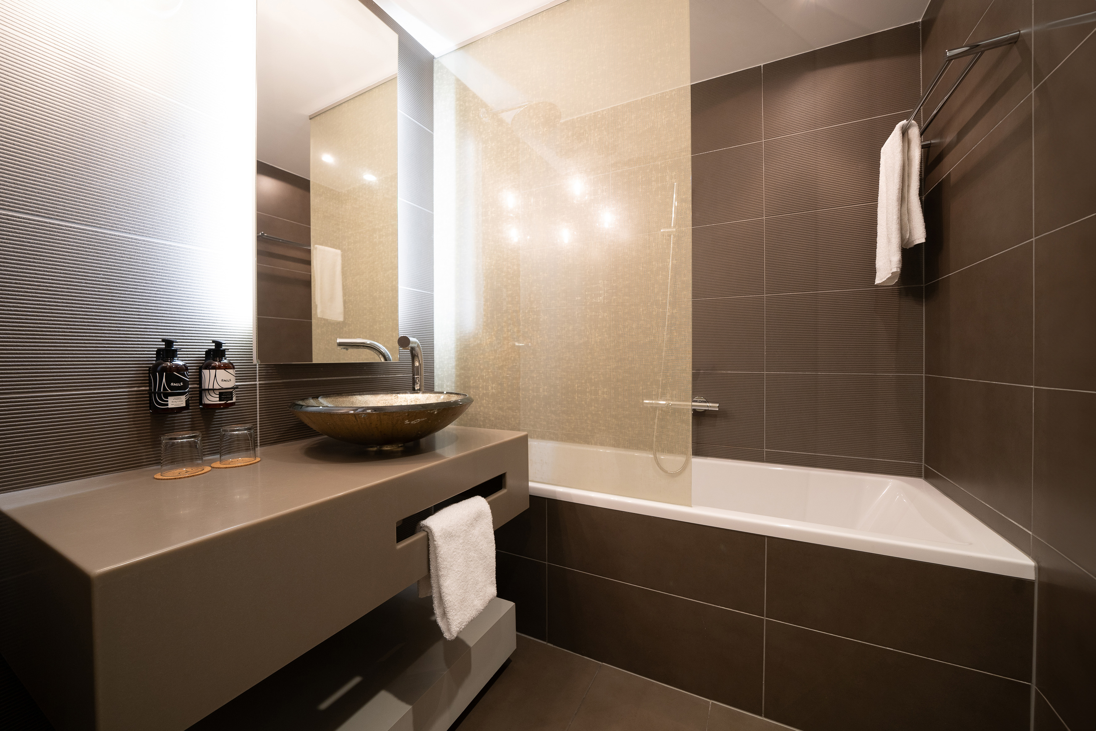 Modern brown-tiled bathroom with floating vanity, glass vessel sink, and bathtub.