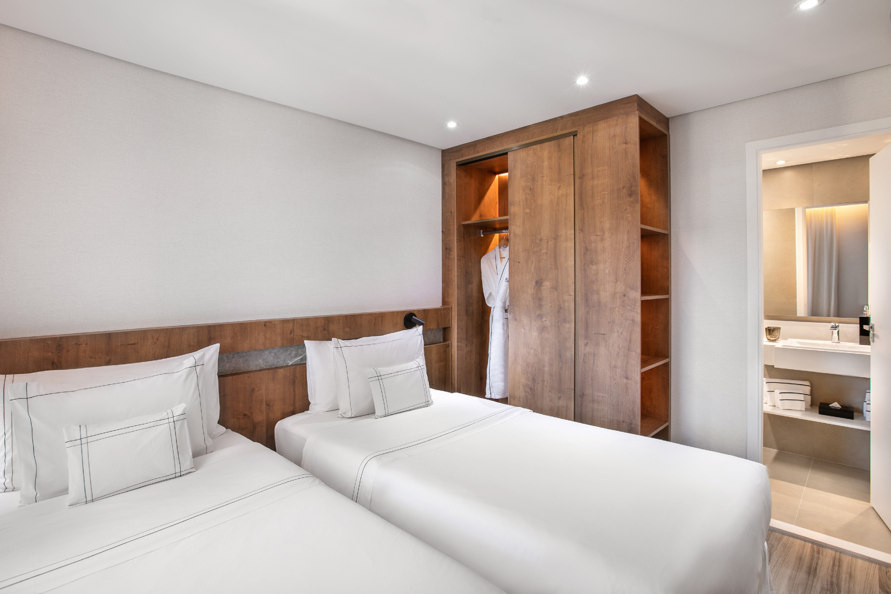 Modern hotel room with two twin beds, wooden wardrobe, and adjoining bathroom.
