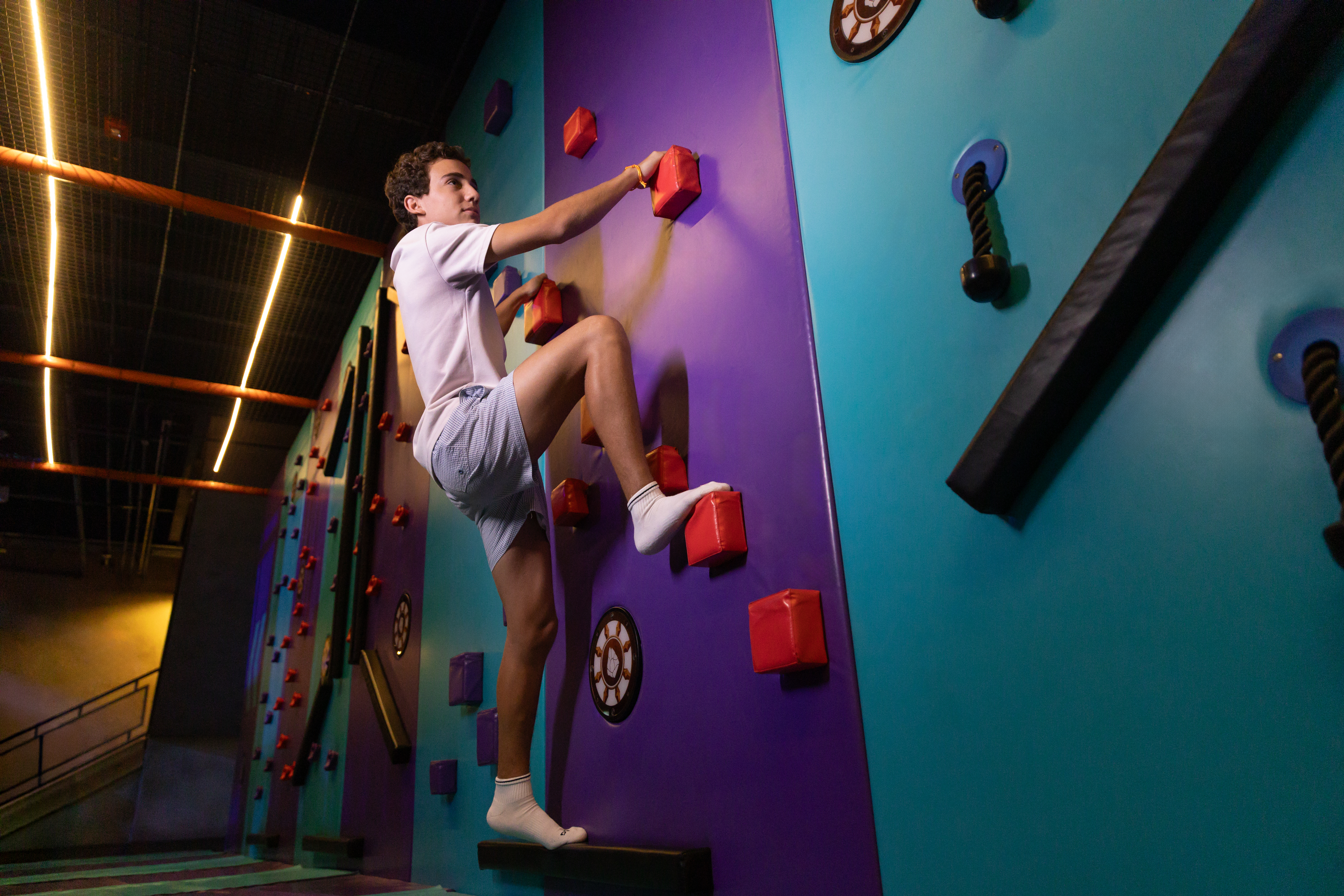 Young man climbing purple and teal indoor wall with red handholds.