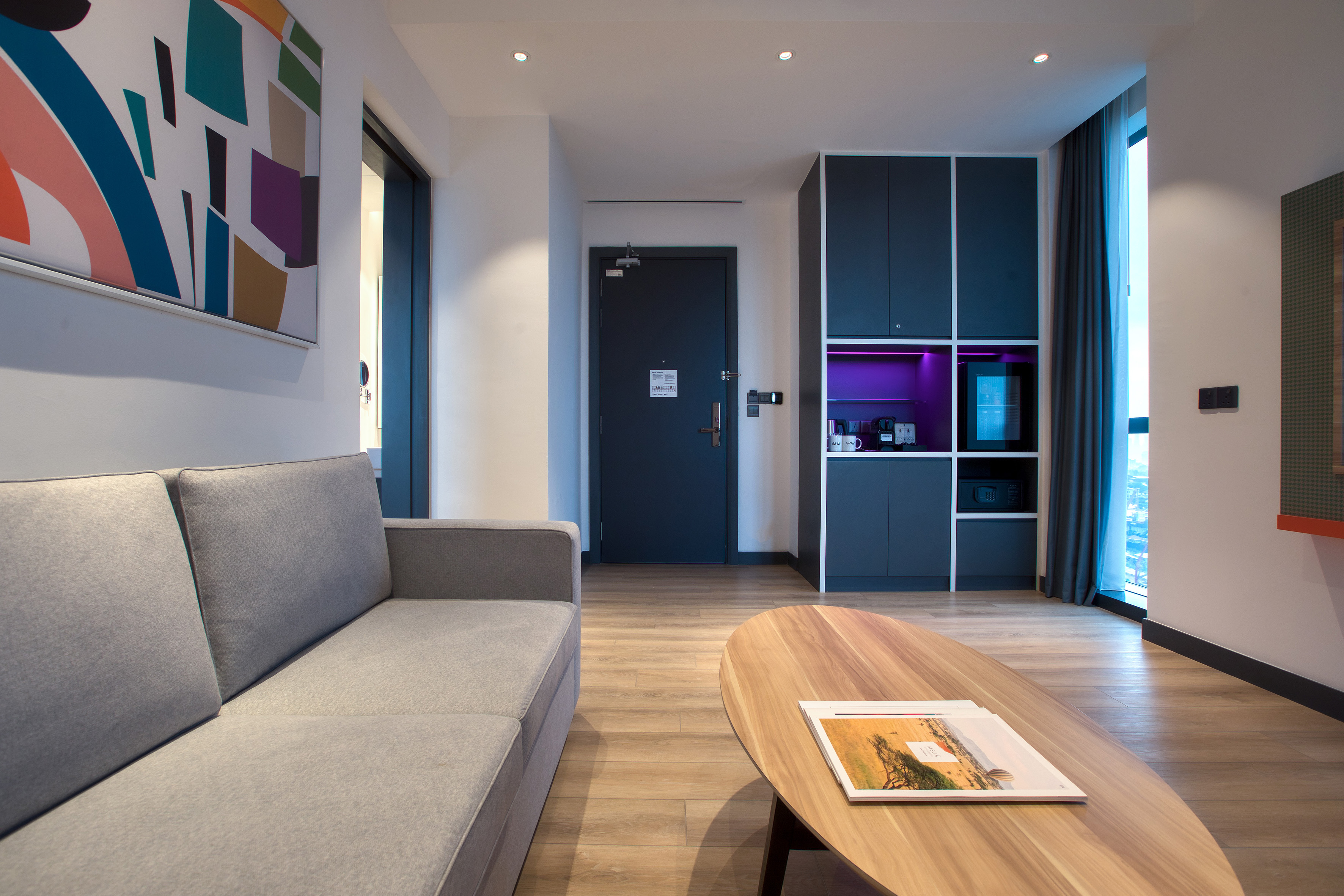 Hotel suite living room with grey sofa, wooden table, abstract art, and mini bar.