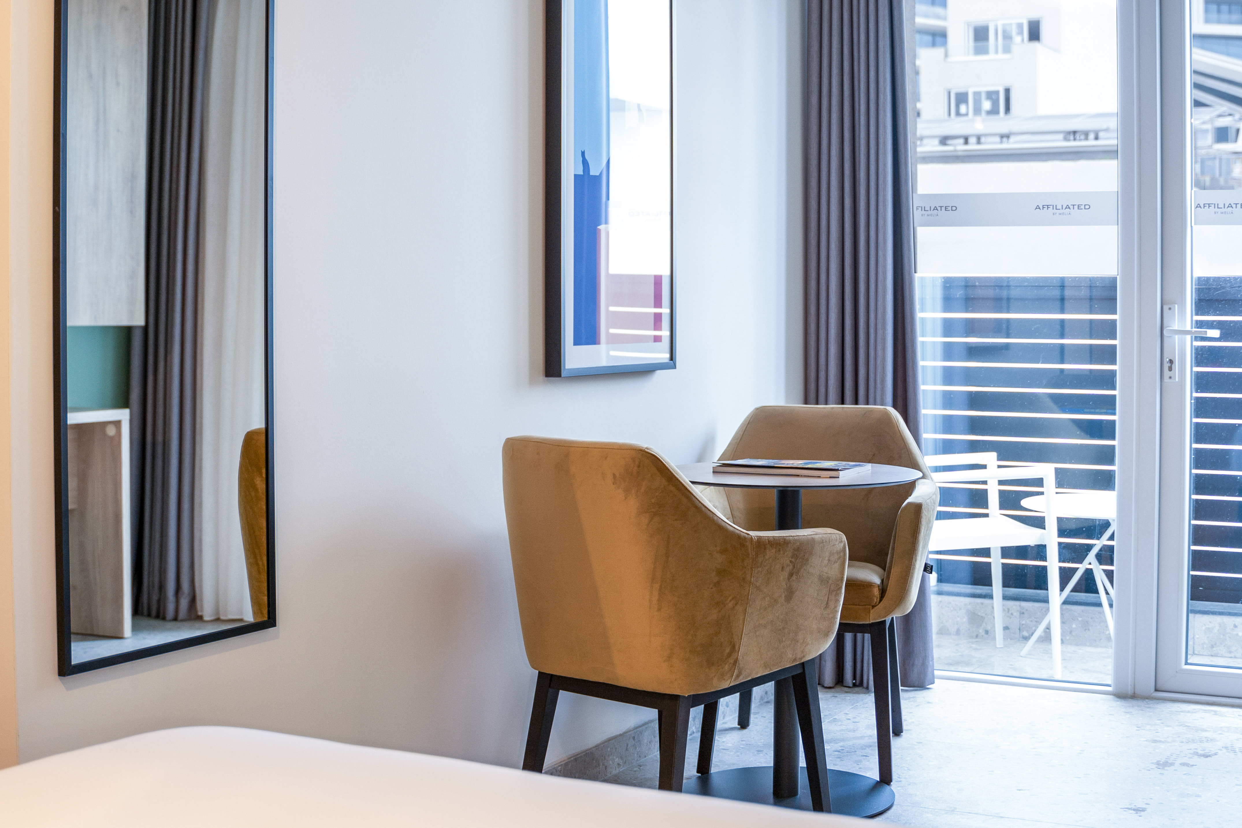 Modern hotel room with two velvet chairs, small table, and balcony access.