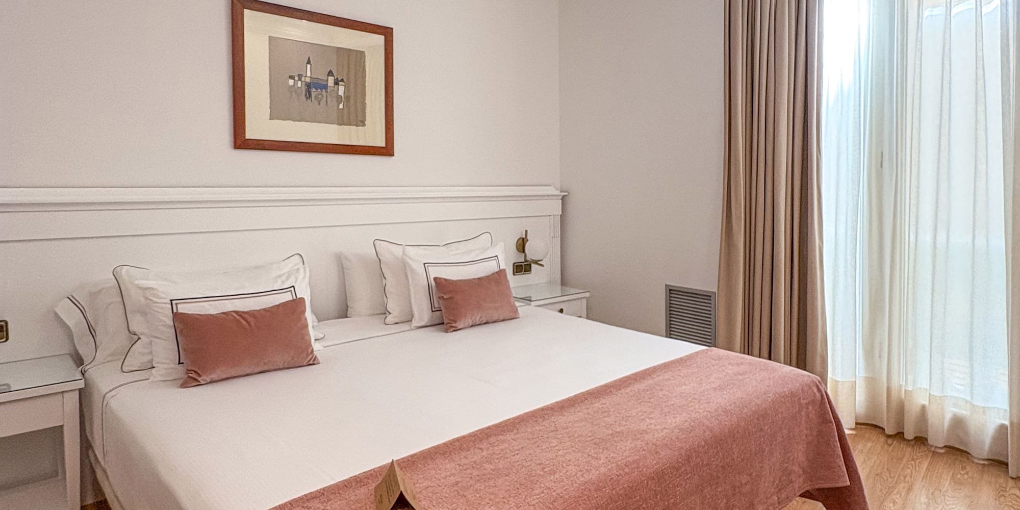 Hotel bedroom with bed, white bedding, pink throw, wooden floor, and bright window.