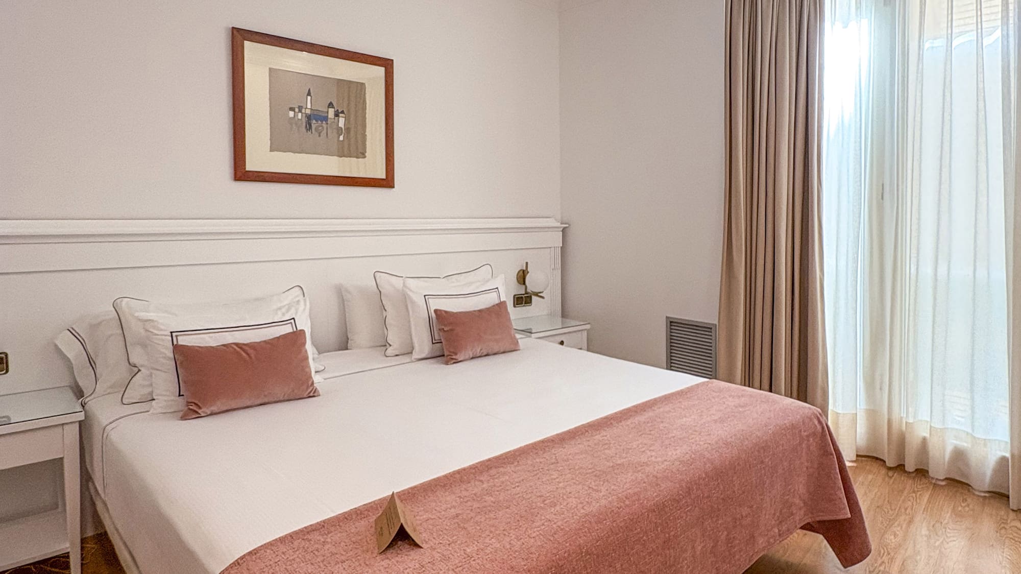Hotel bedroom with bed, white bedding, pink throw, wooden floor, and bright window.