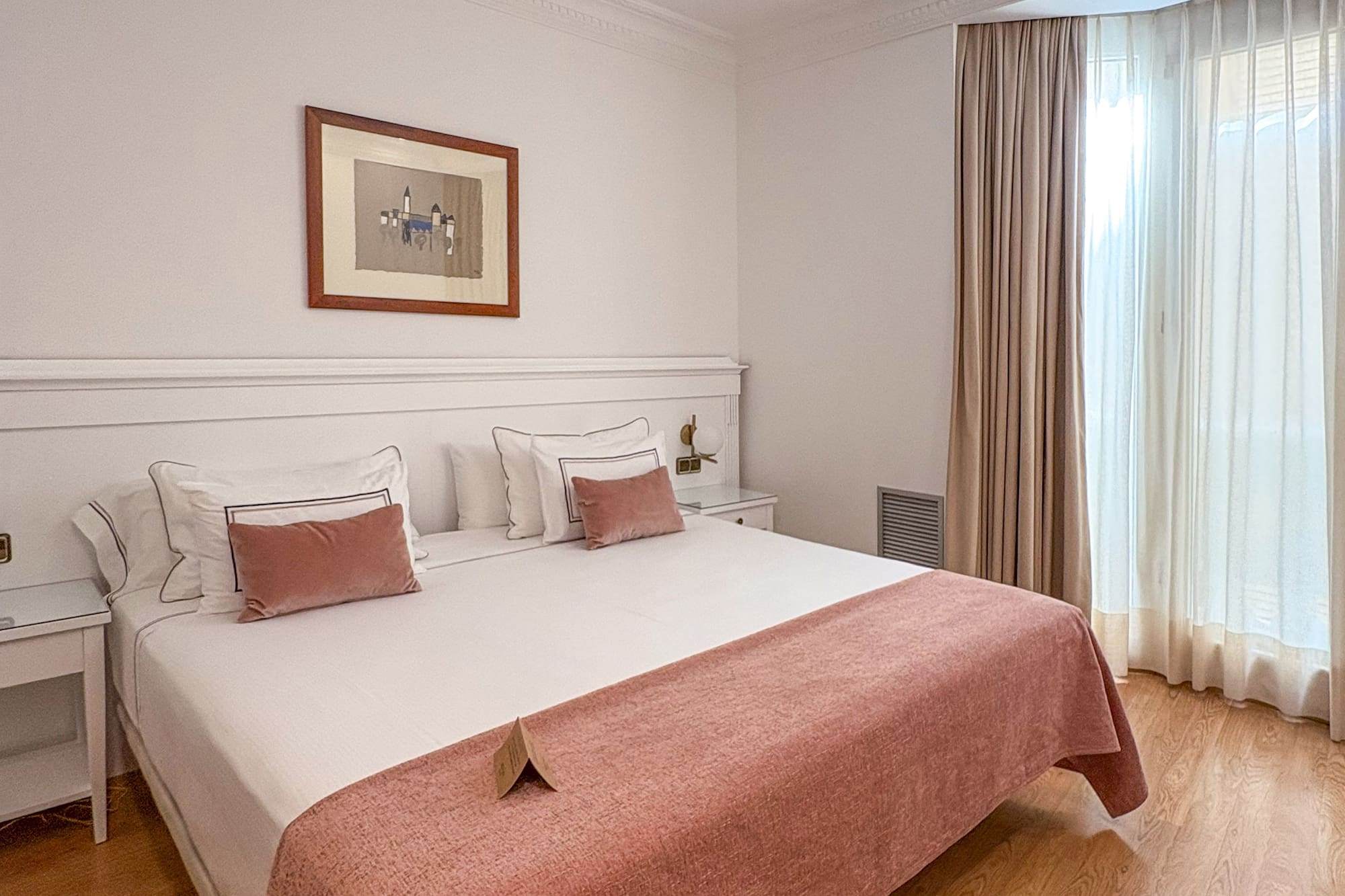 Hotel bedroom with bed, white bedding, pink throw, wooden floor, and bright window.