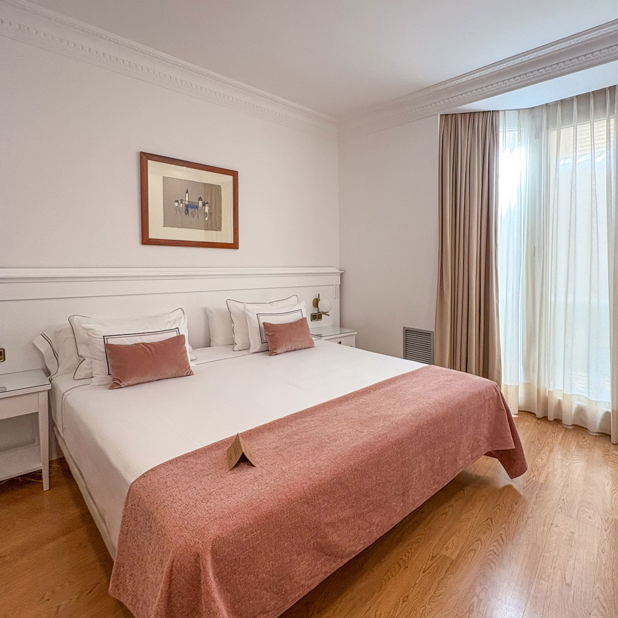 Hotel bedroom with bed, white bedding, pink throw, wooden floor, and bright window.