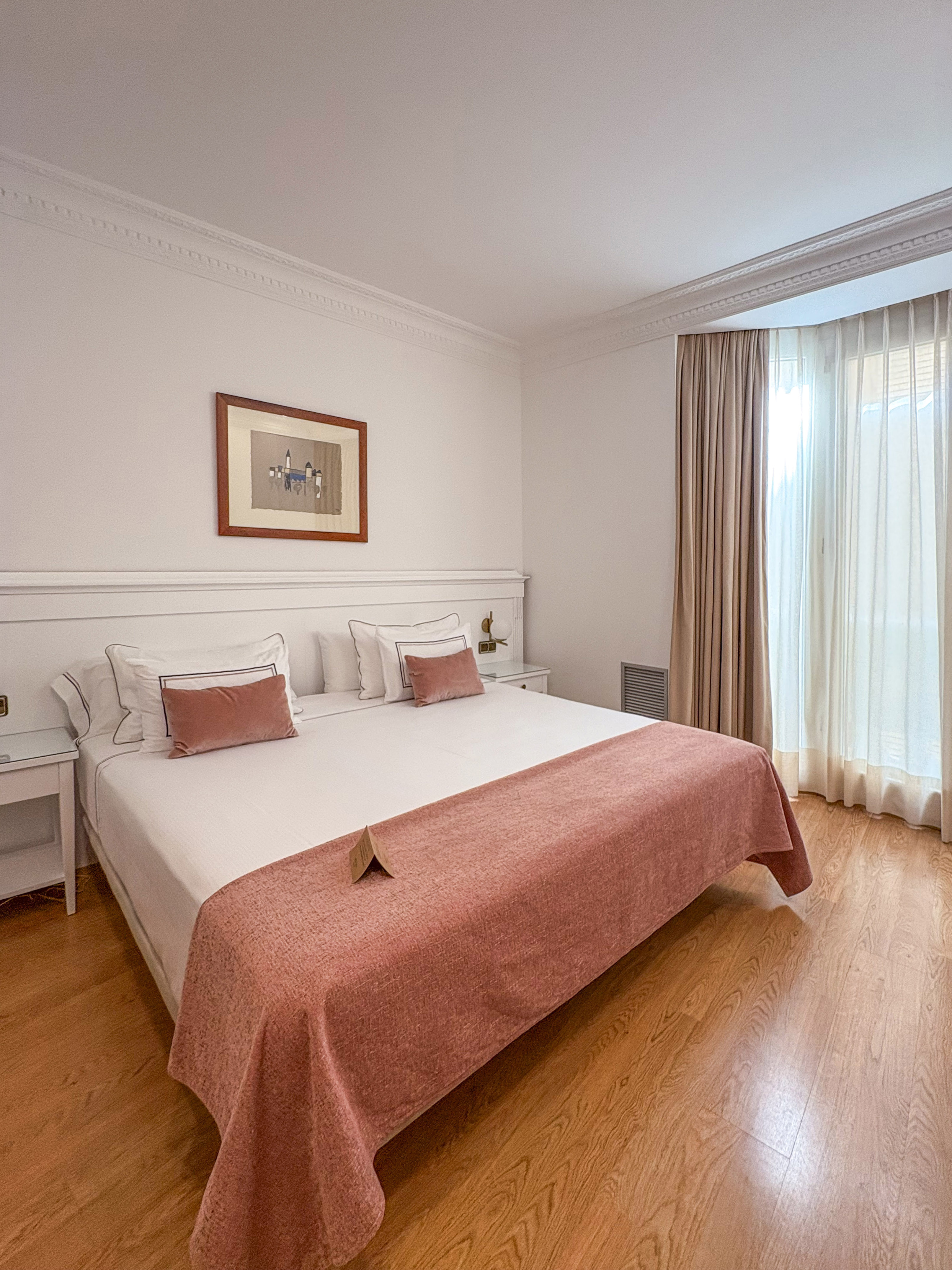 Hotel bedroom with bed, white bedding, pink throw, wooden floor, and bright window.