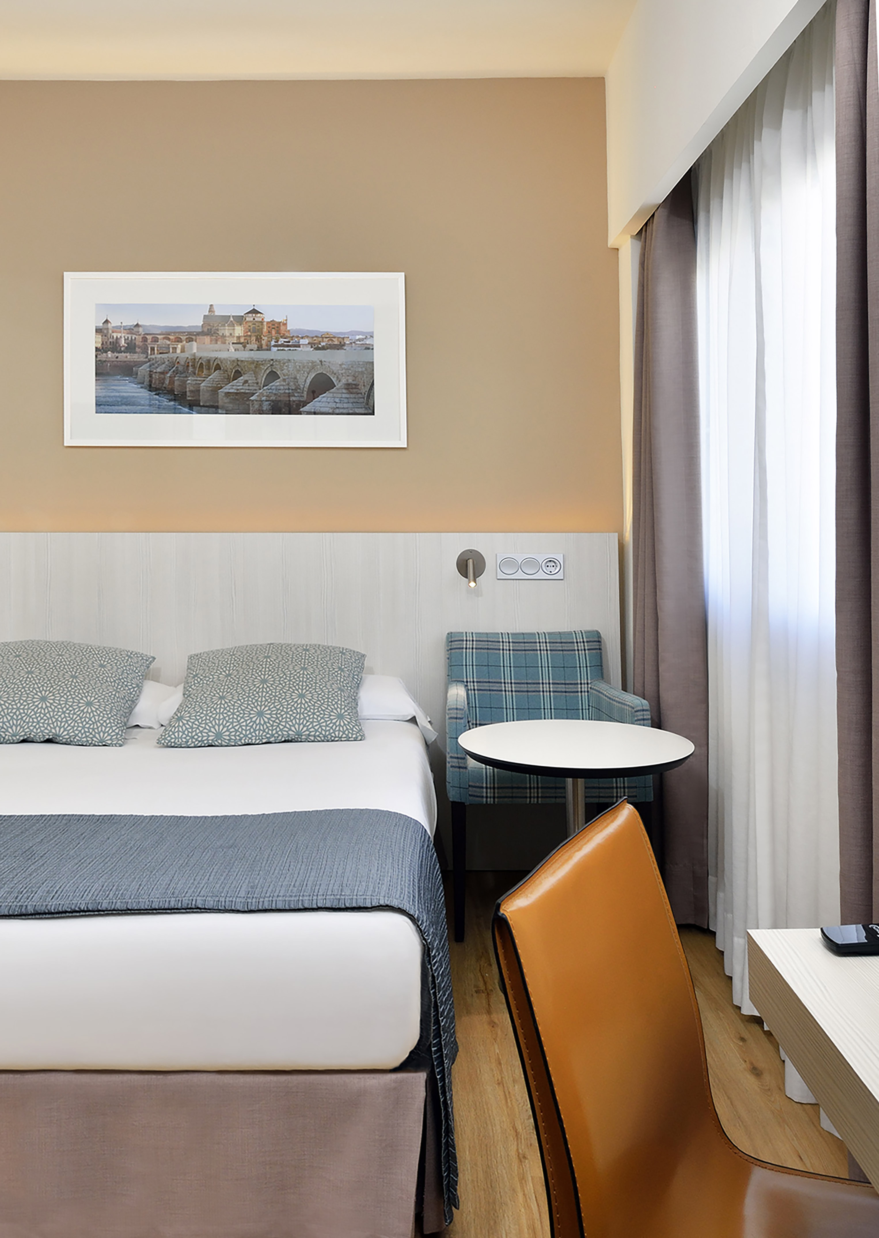 Modern hotel room with bed, Cordoba bridge art, plaid armchair, and brown chair.