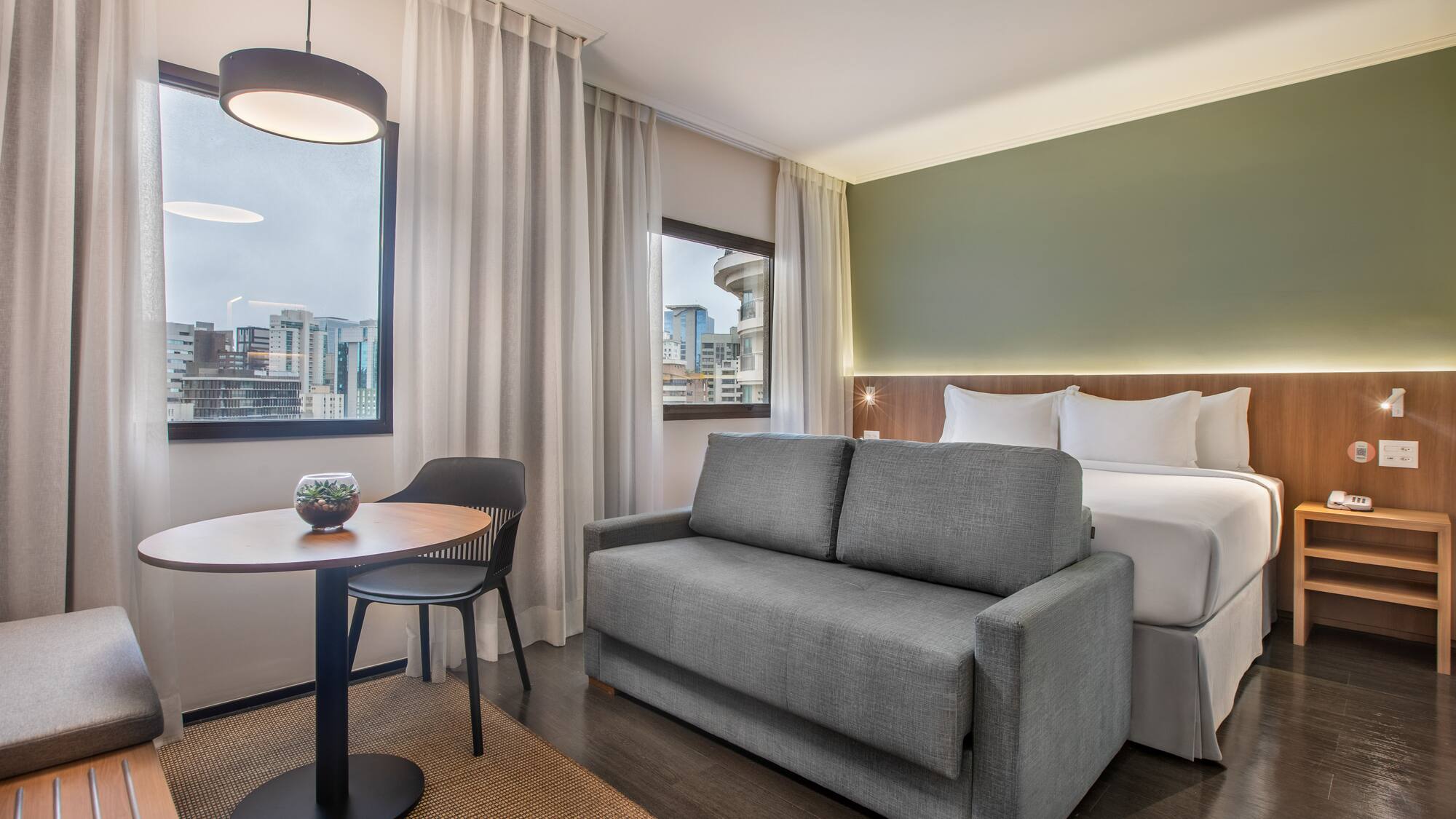 Modern hotel room with bed, grey sofa, dining table, and cityscape view.