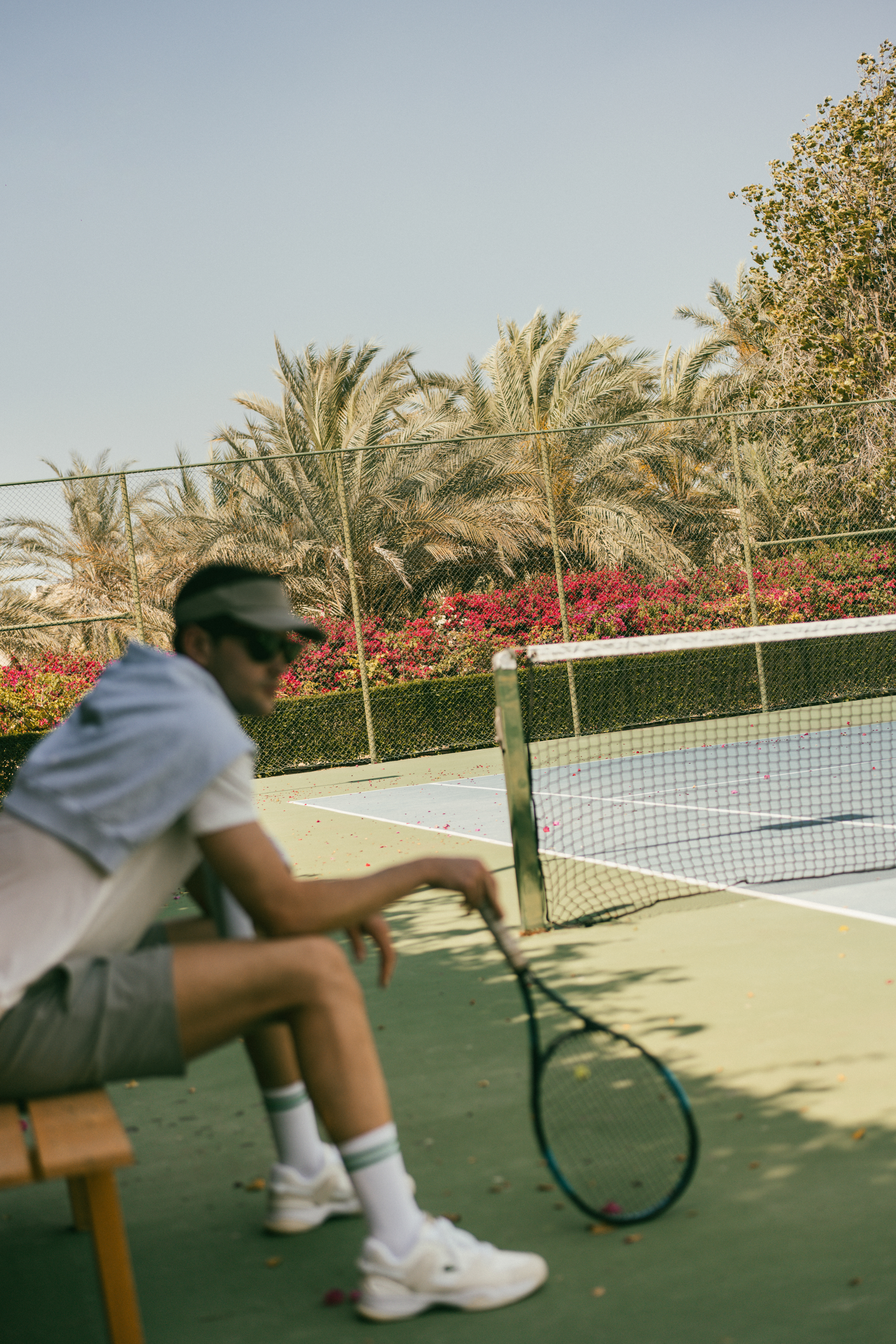 Tennis player with racket resting on court bench under palm trees.