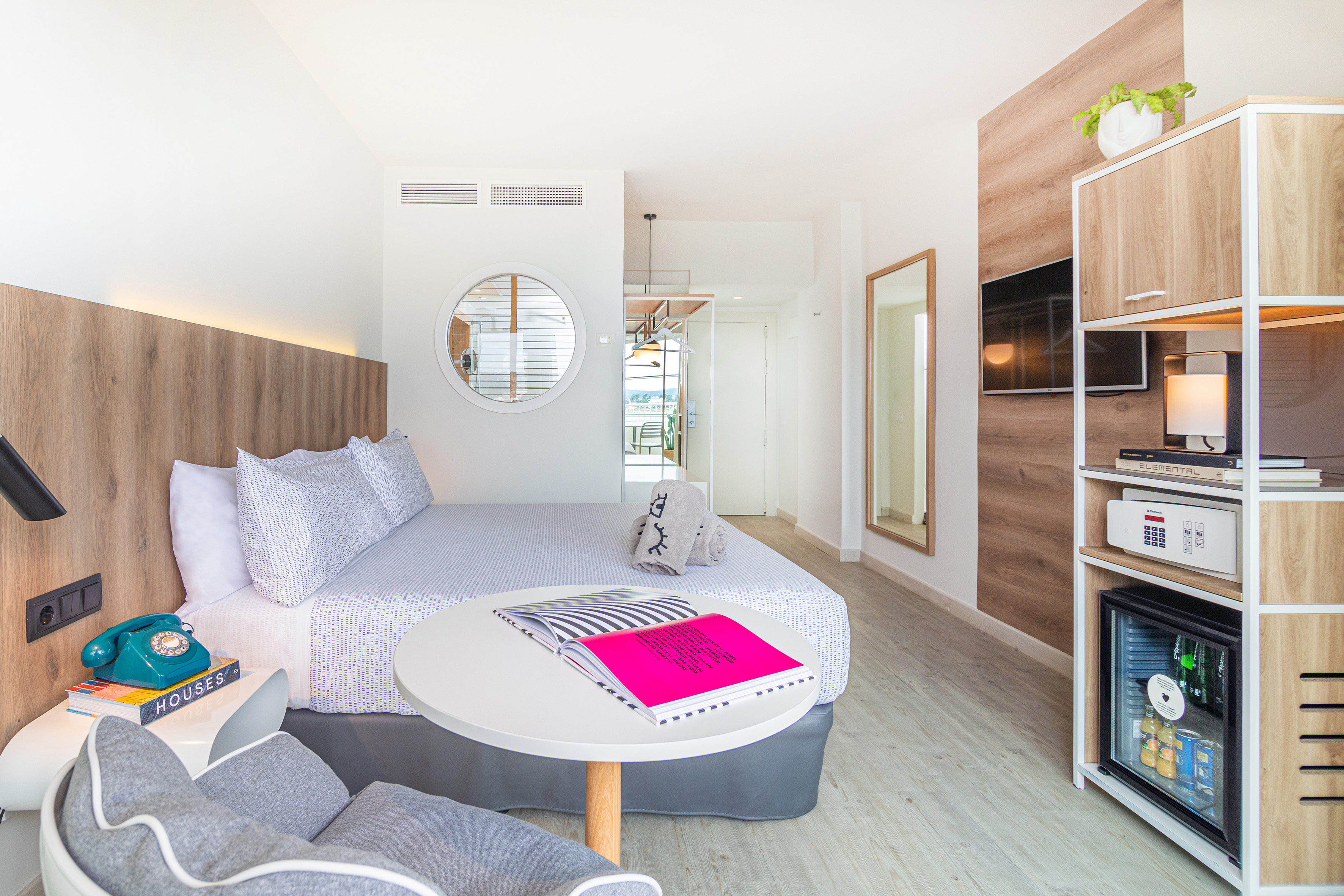 Modern hotel room with bed, wooden wall, TV, mini-fridge, and mirrors.