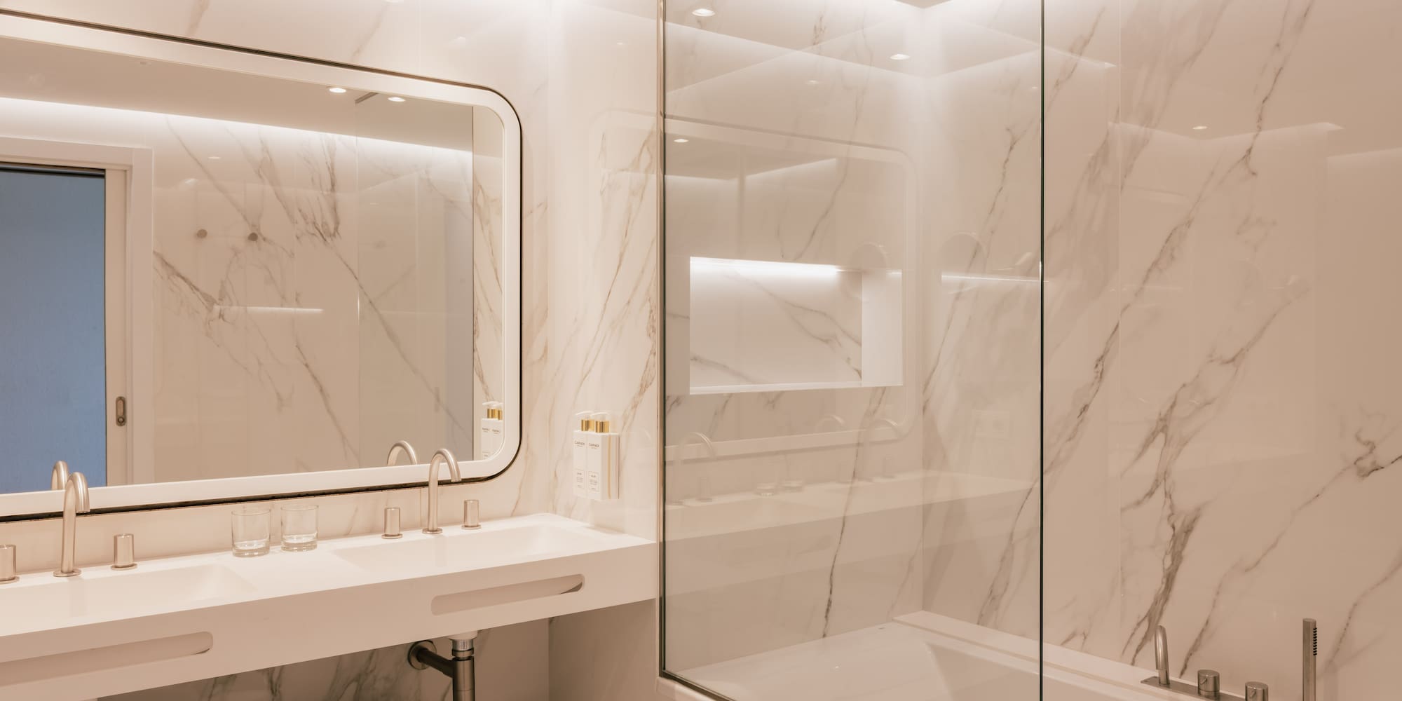 Modern marble bathroom with double vanity, glass shower-tub, and white towels.