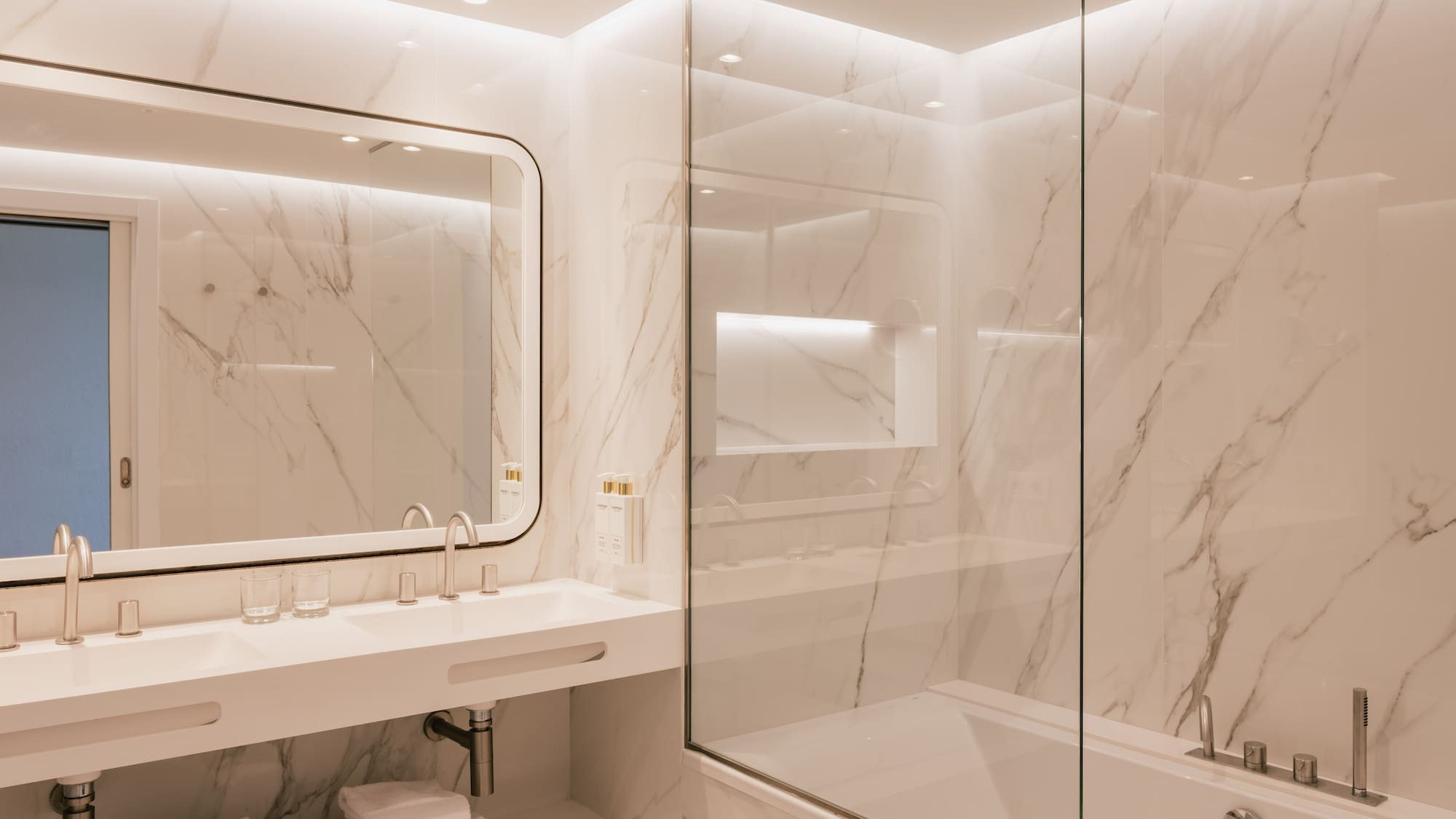 Modern marble bathroom with double vanity, glass shower-tub, and white towels.