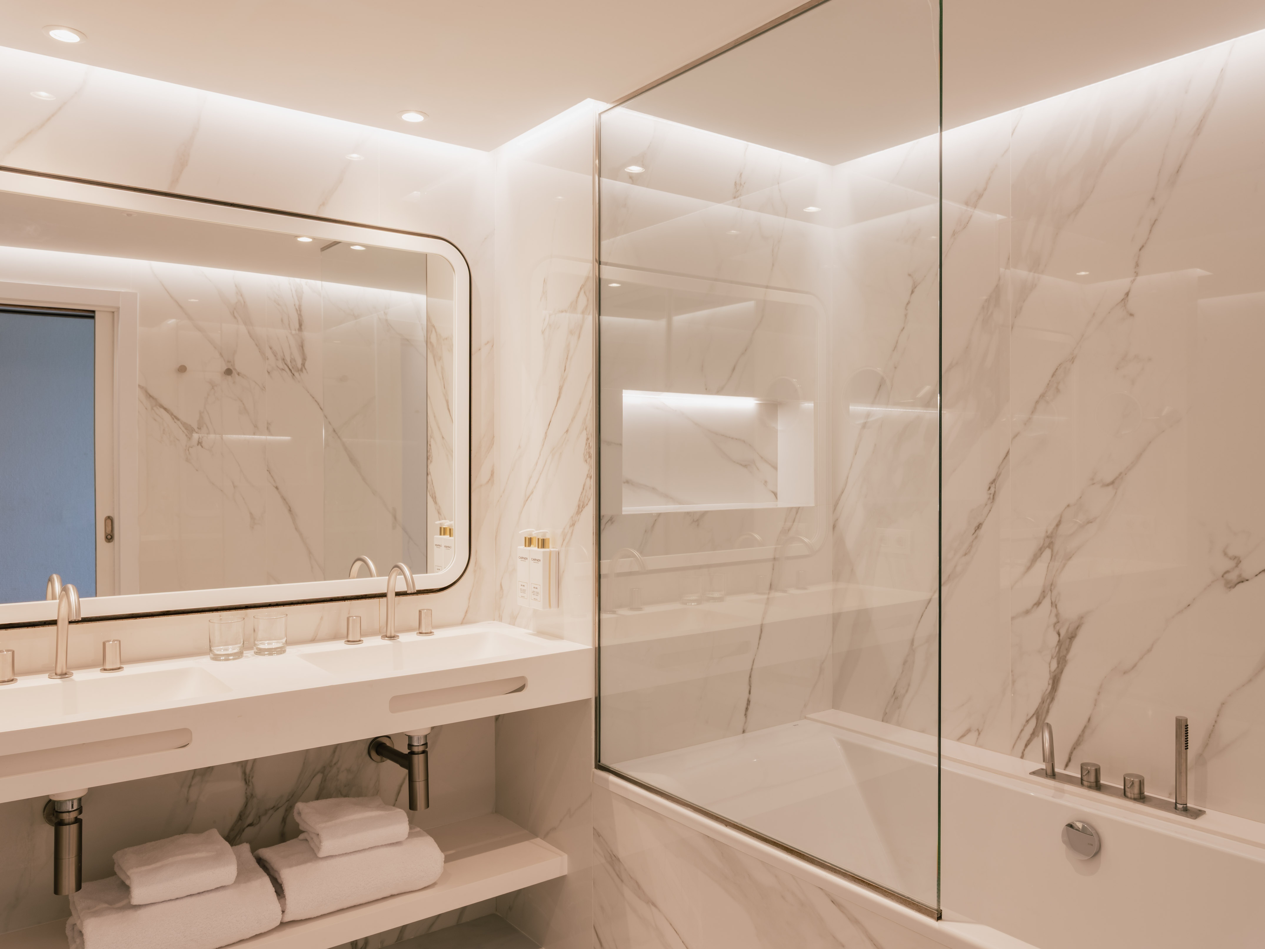 Baño minimalista de mármol blanco con doble lavabo, bañera y ducha de vidrio.