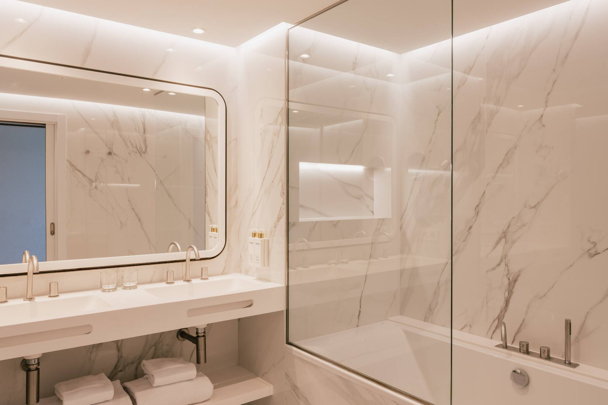 Baño minimalista de mármol blanco con doble lavabo, bañera y ducha de vidrio.