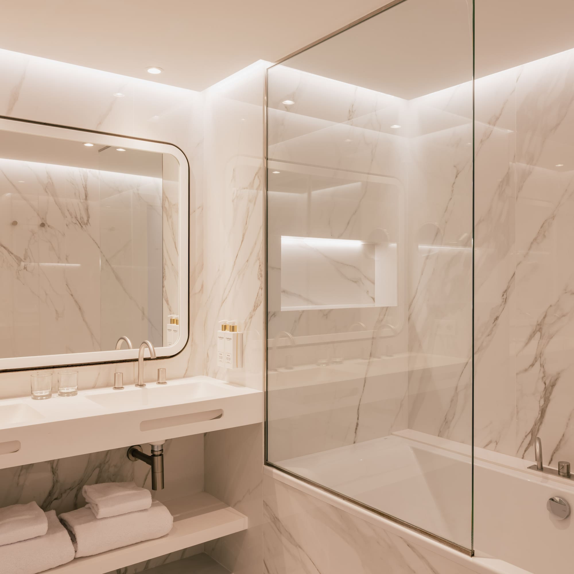 Modern marble bathroom with double vanity, glass shower-tub, and white towels.