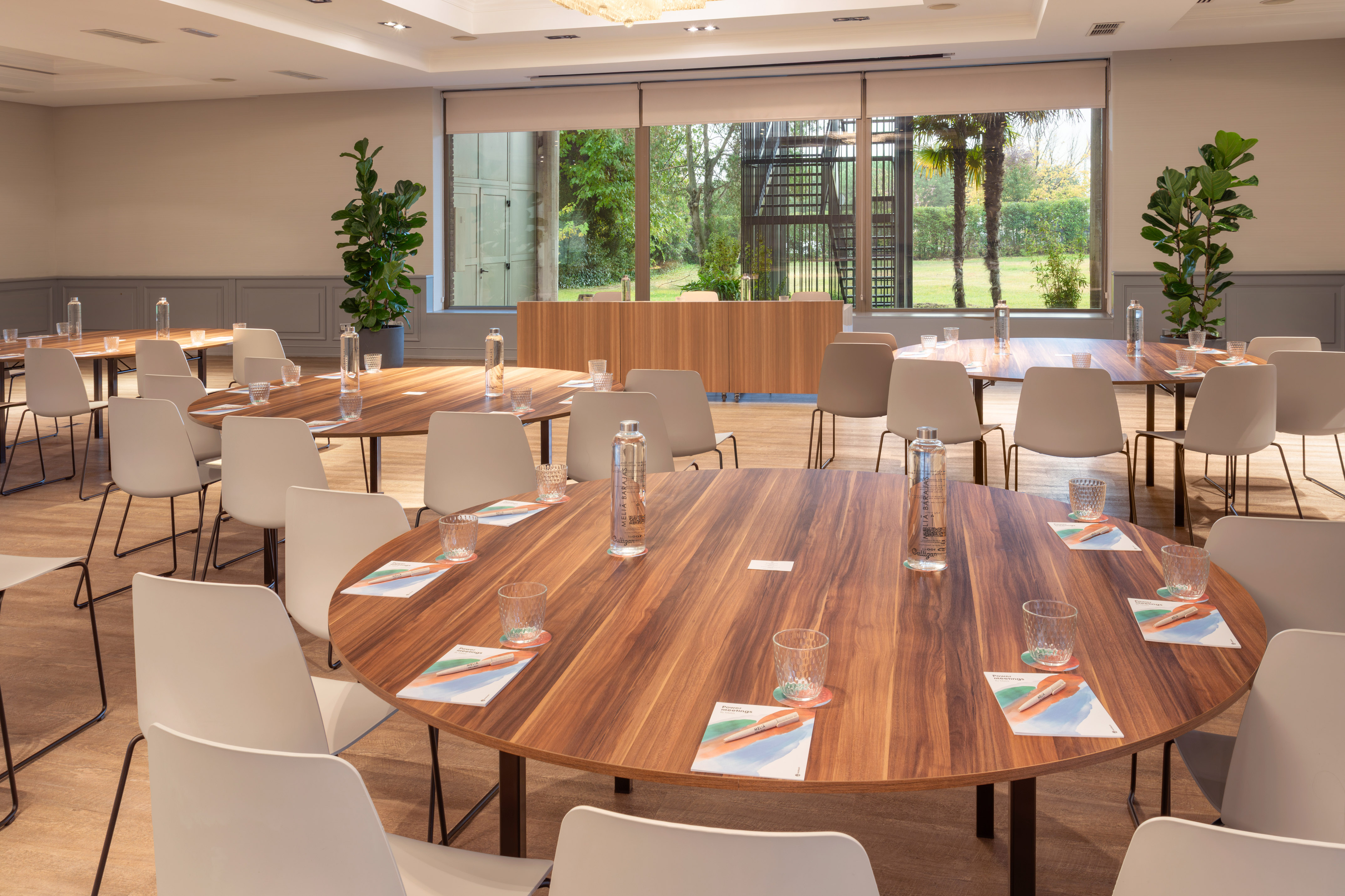 Modern conference room with round wooden tables, white chairs, and garden view.