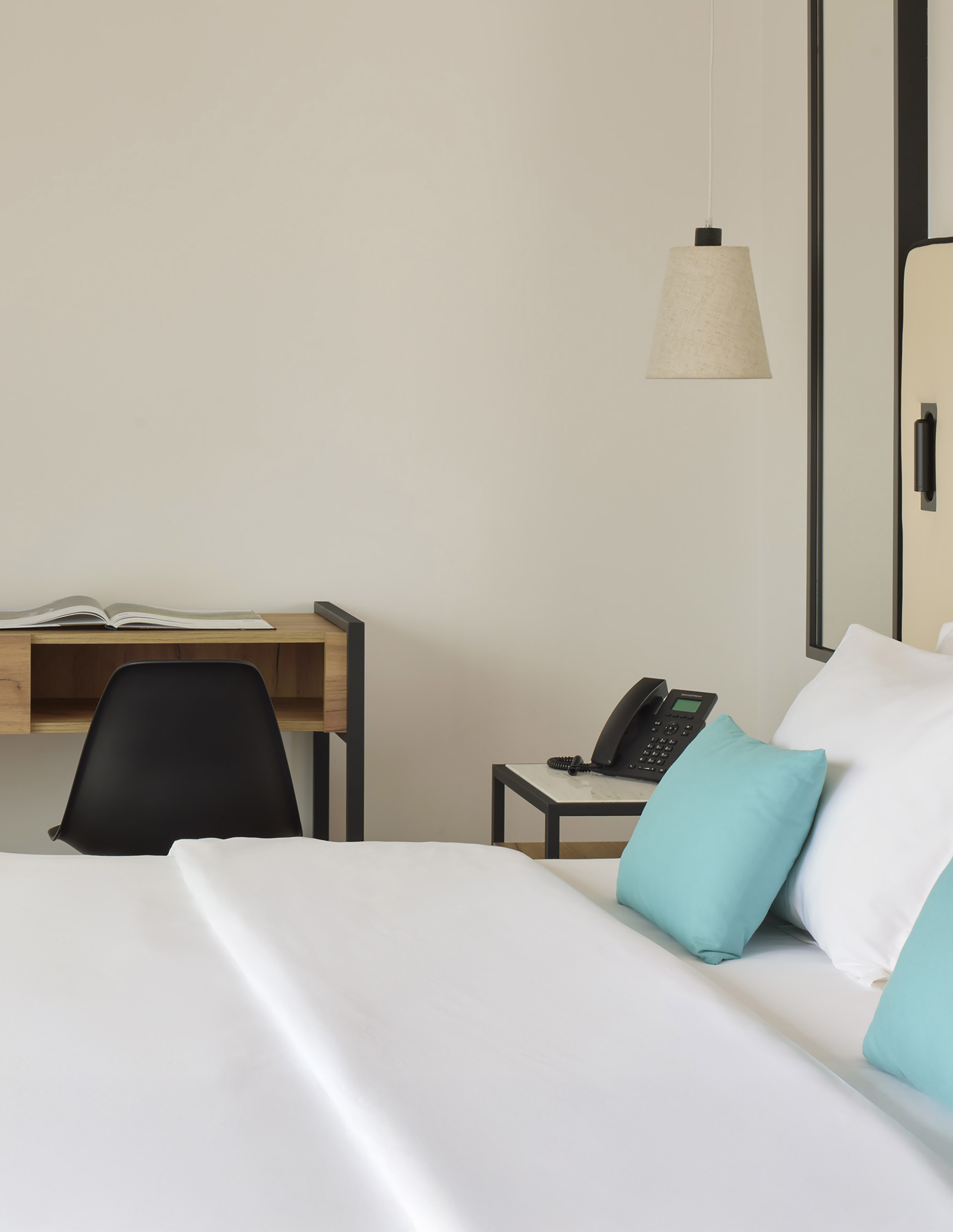 Modern hotel bedroom with white bed, teal pillows, wooden desk, and phone.
