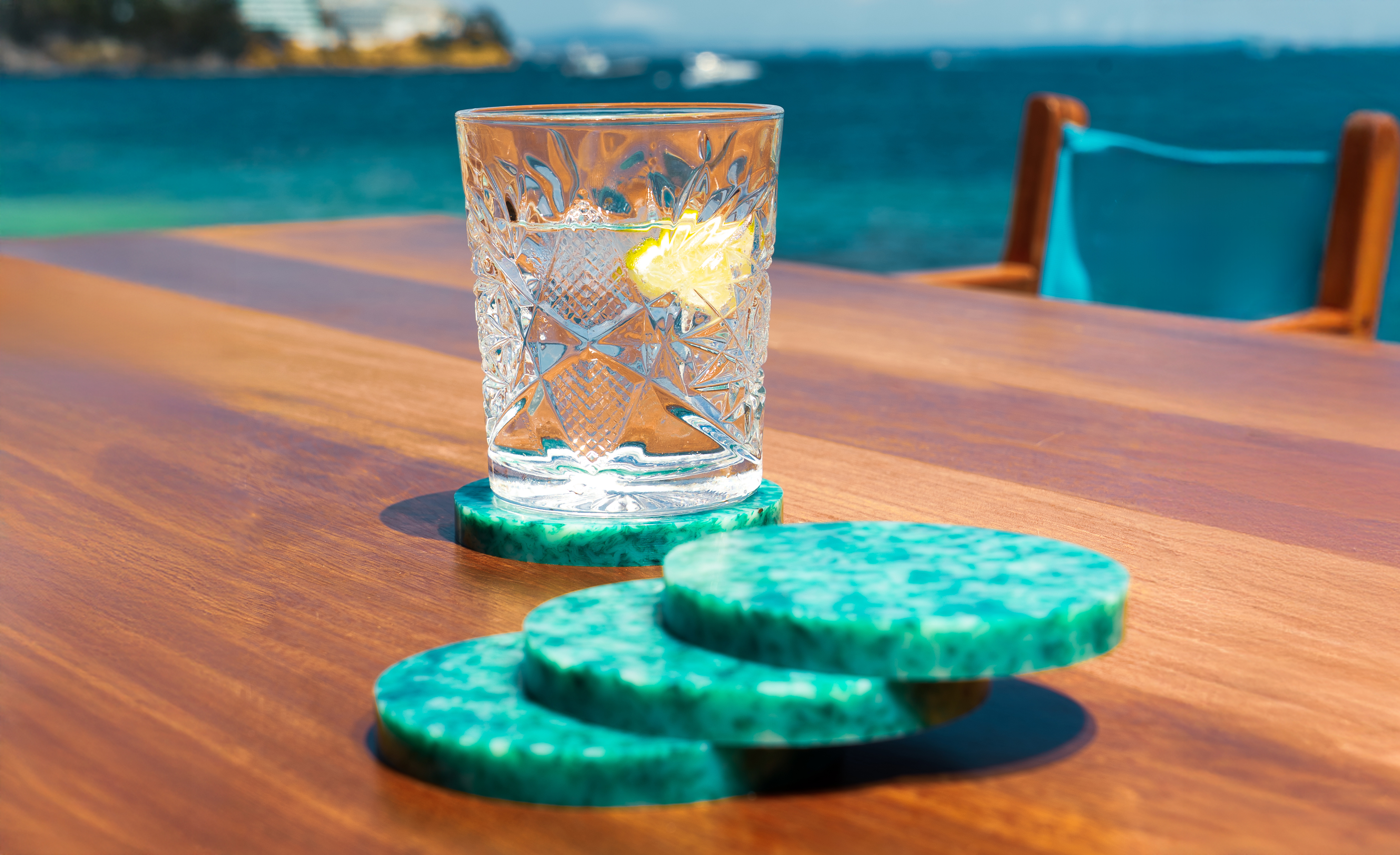 Glass of water, lemon, and green patterned coasters on wooden table by the sea.