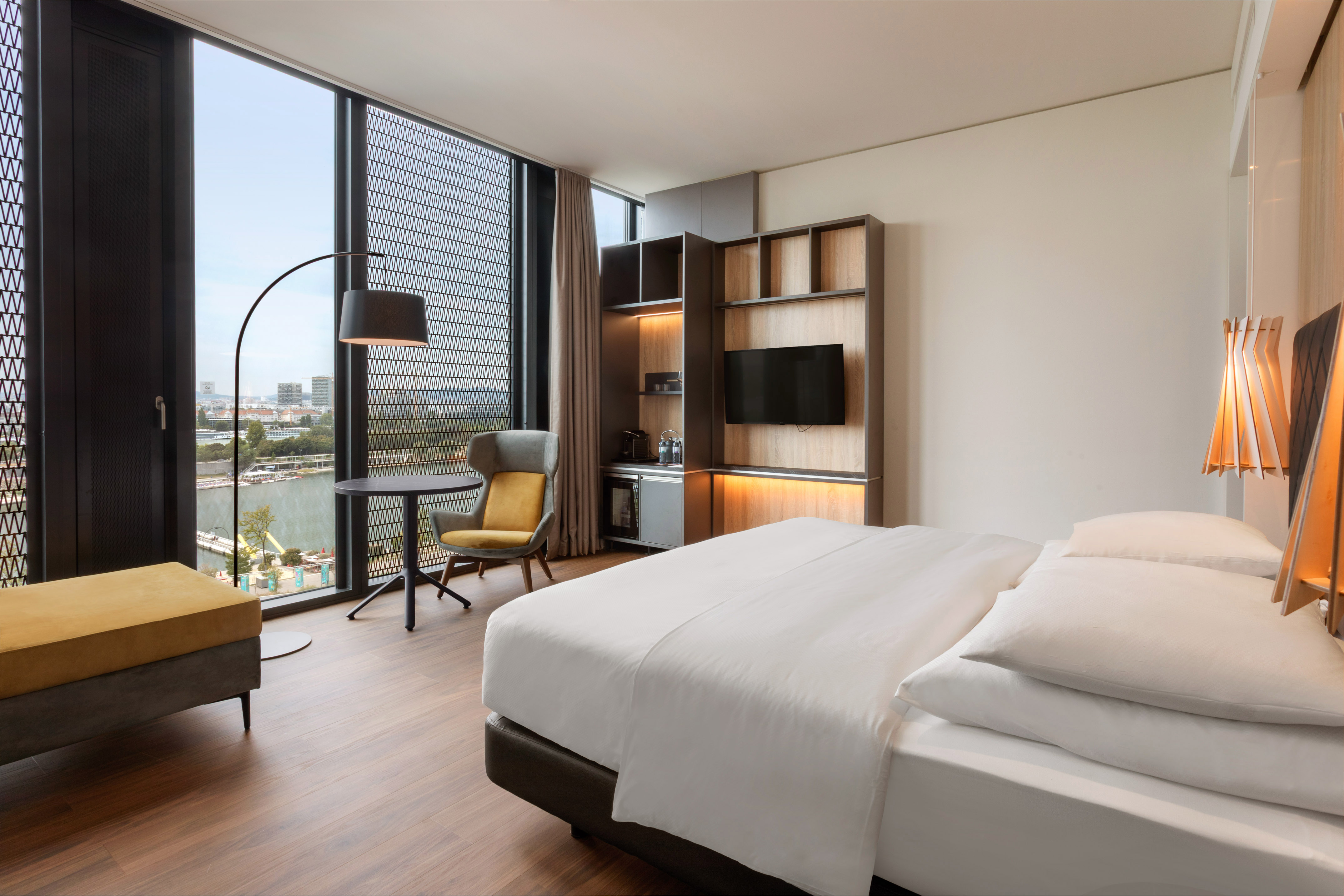 Modern hotel room with bed, large window waterfront view, armchair, and TV.