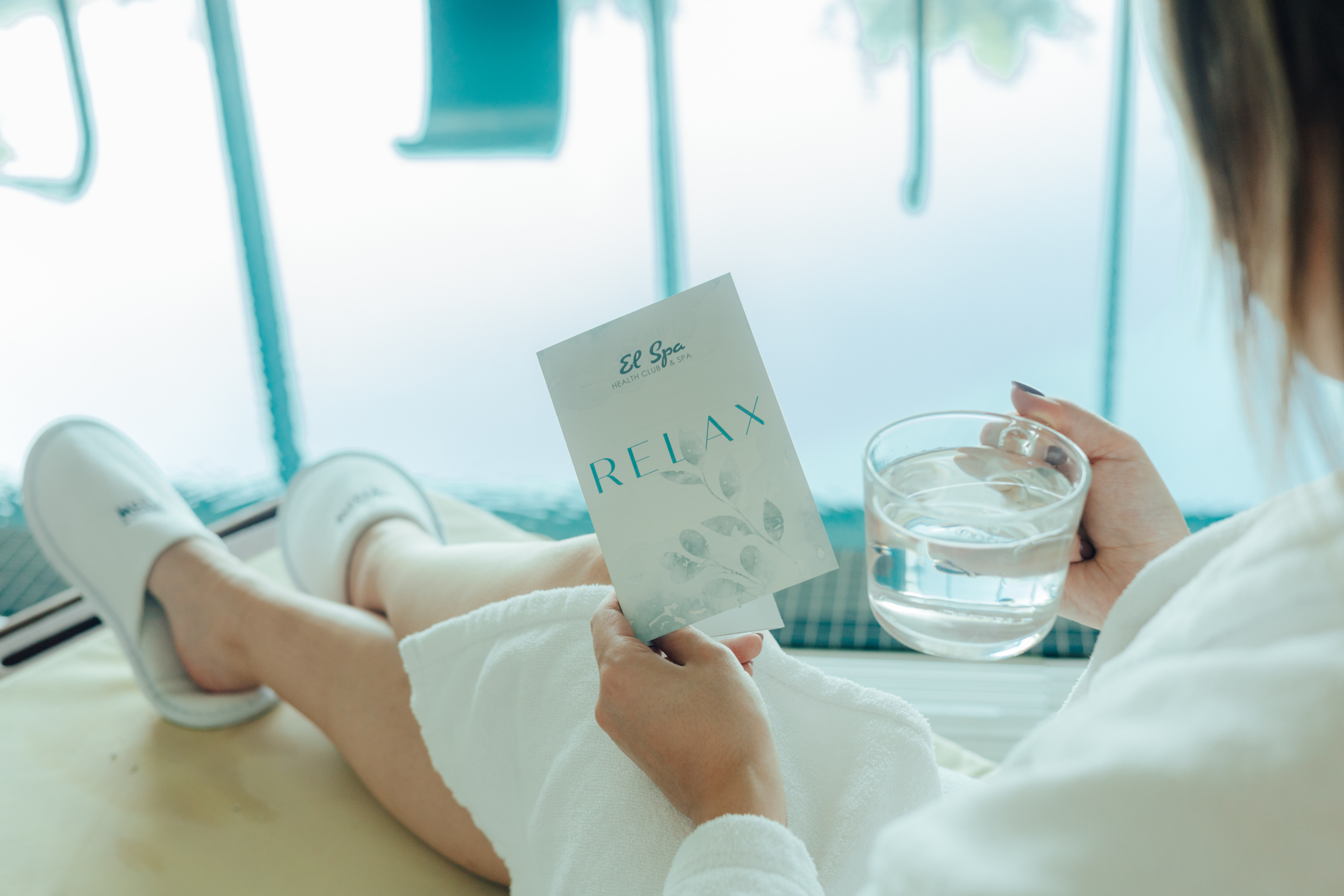 Woman in spa robe holding "El Spa Relax" brochure and water glass.