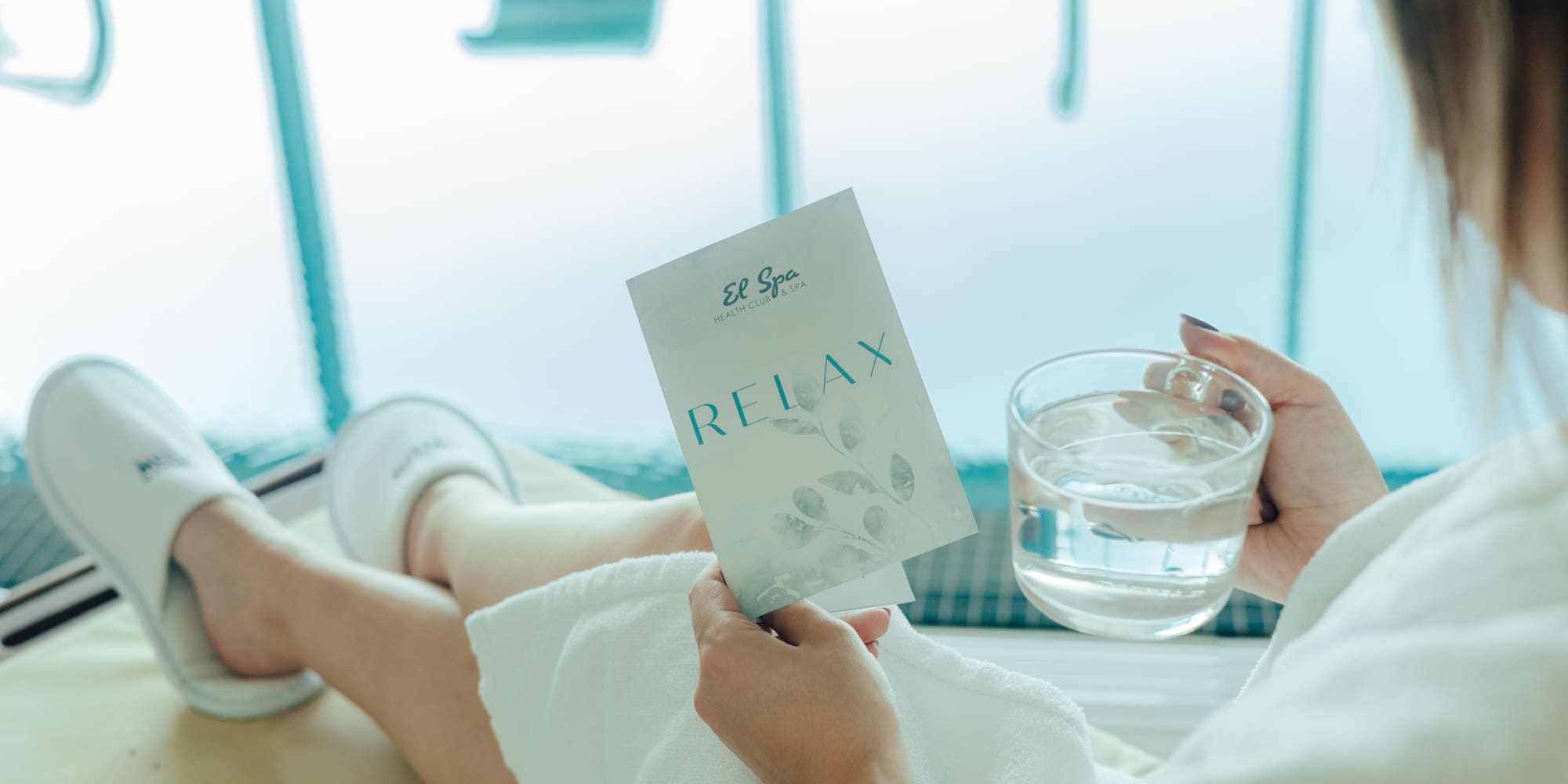 Woman in spa robe holding "El Spa Relax" brochure and water glass.