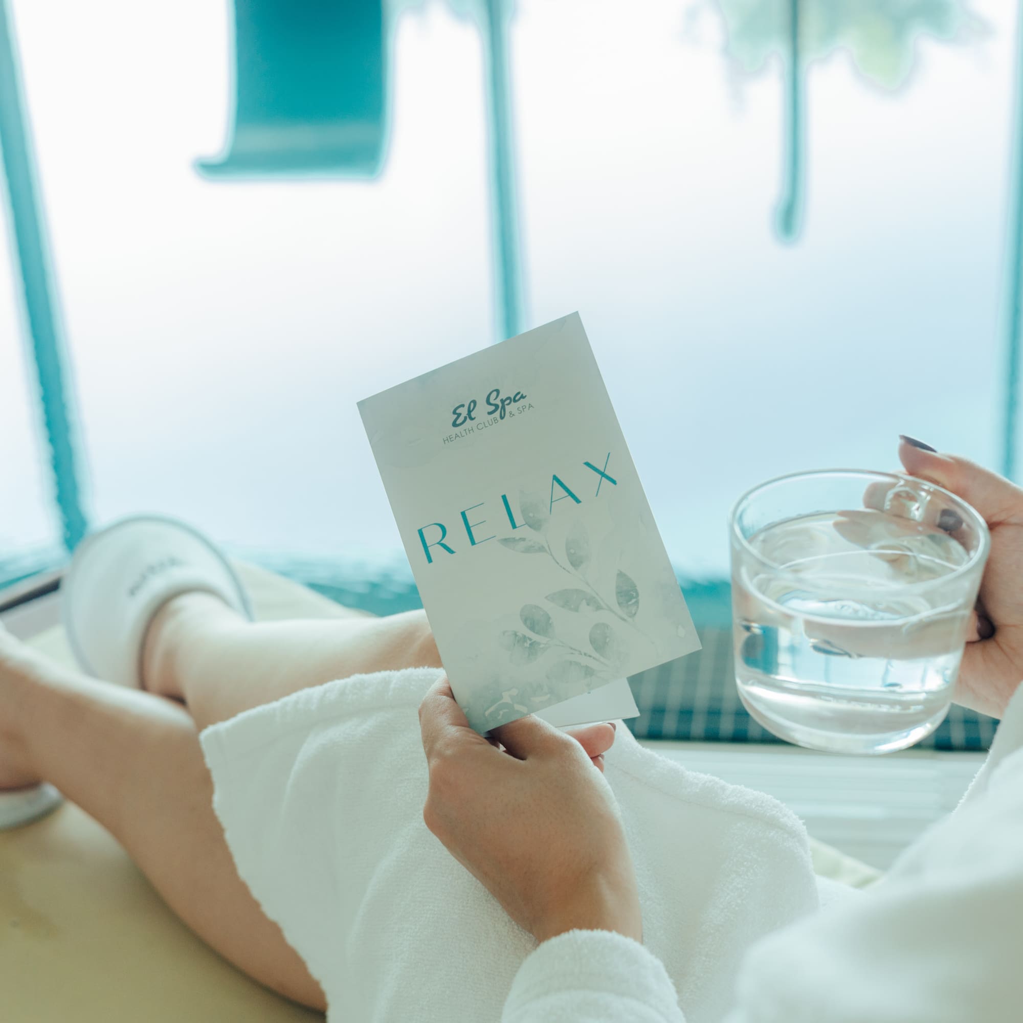 Woman in spa robe holding "El Spa Relax" brochure and water glass.