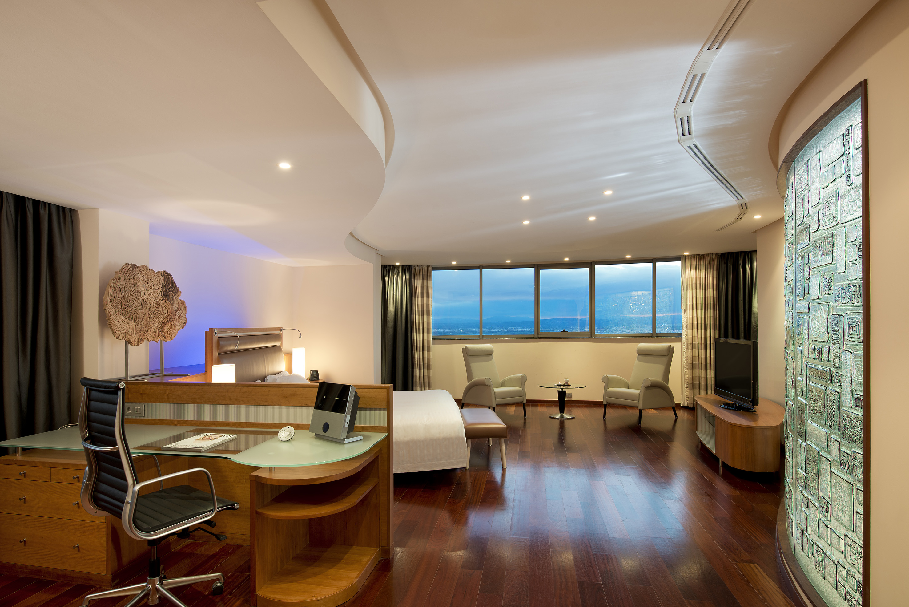 Modern hotel suite with desk, bed, seating, dark wood floor, and city view.