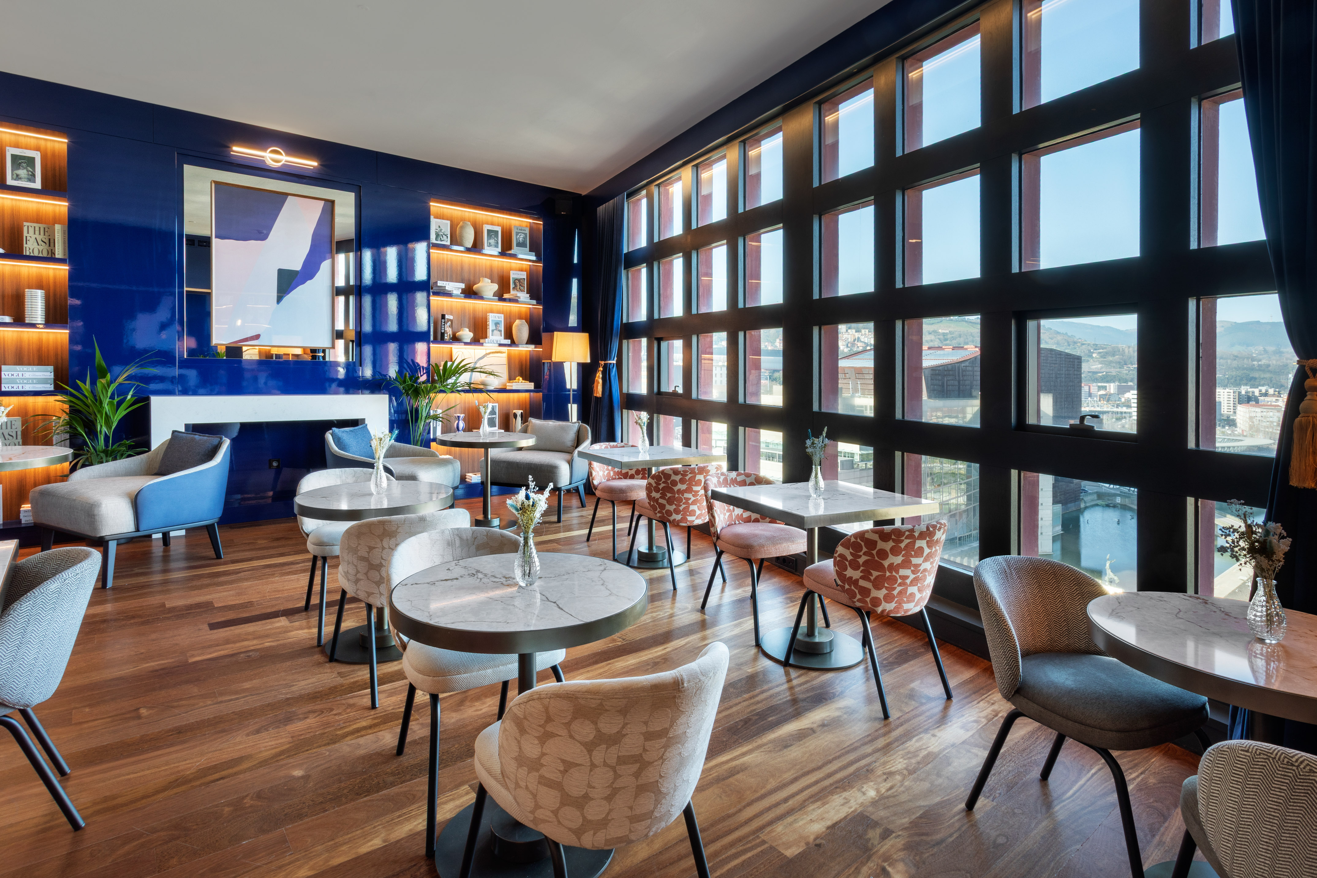Modern hotel lounge with blue walls, stylish seating, marble tables, city view.