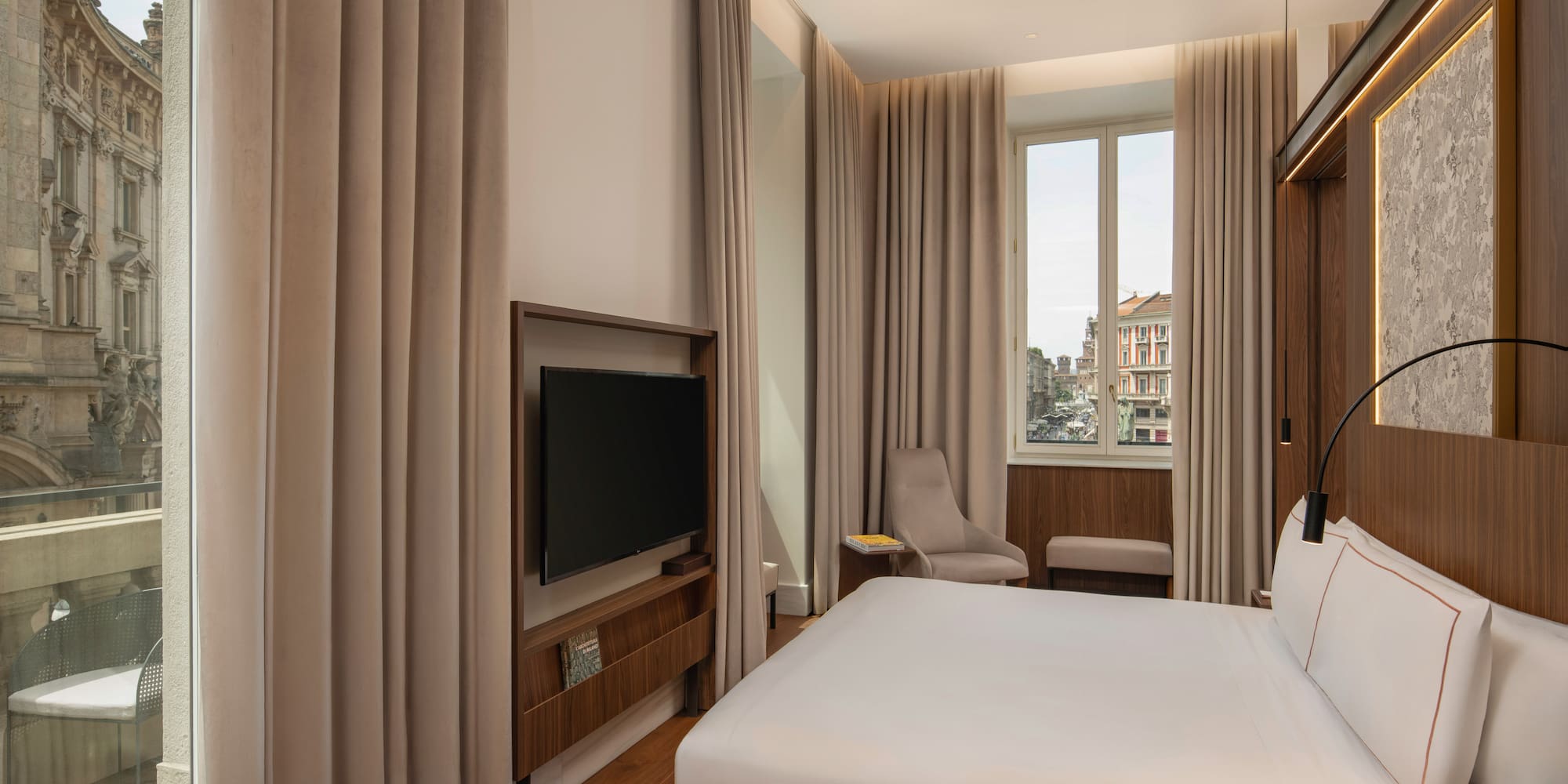 Modern hotel room with king bed, city view, wood floors, and wall-mounted TV.