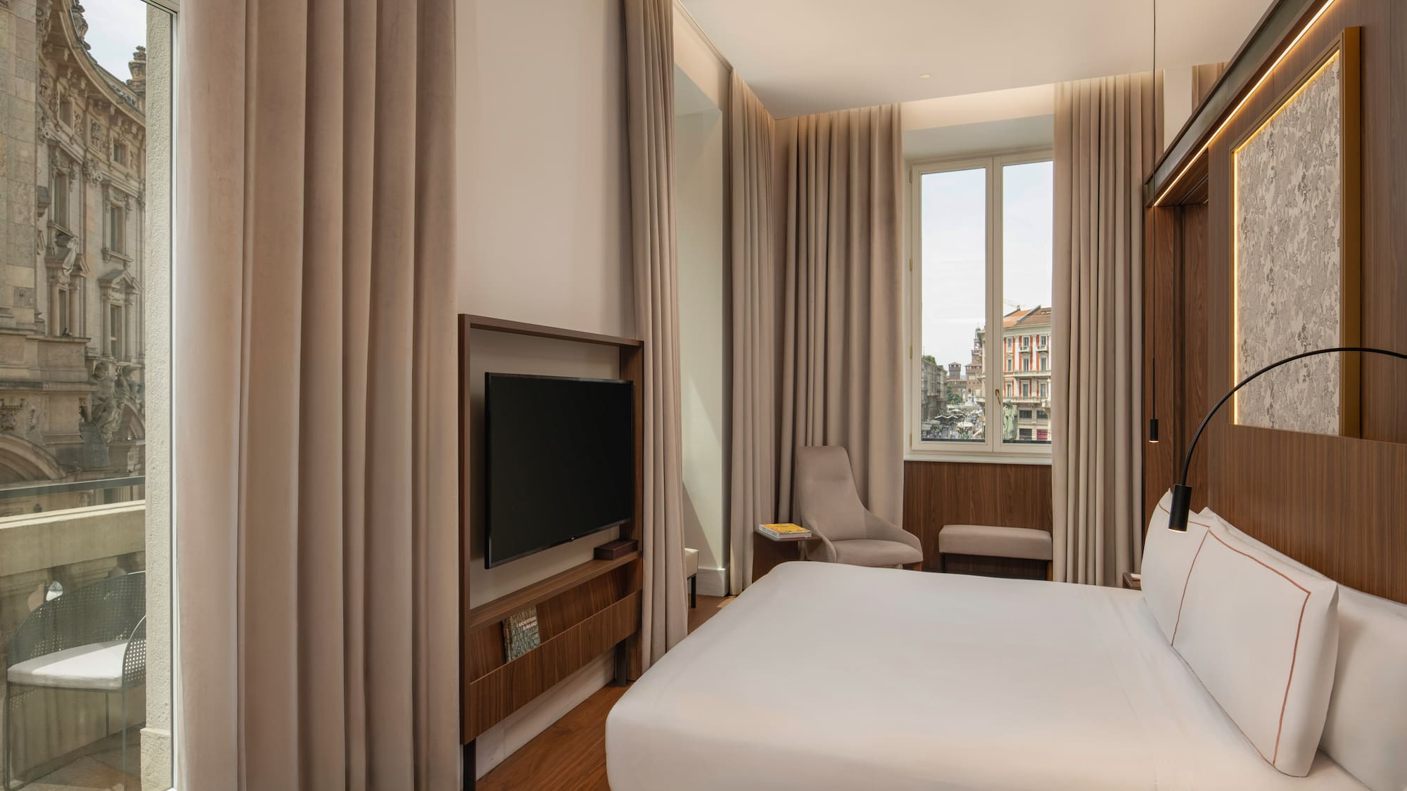 Modern hotel room with king bed, city view, wood floors, and wall-mounted TV.