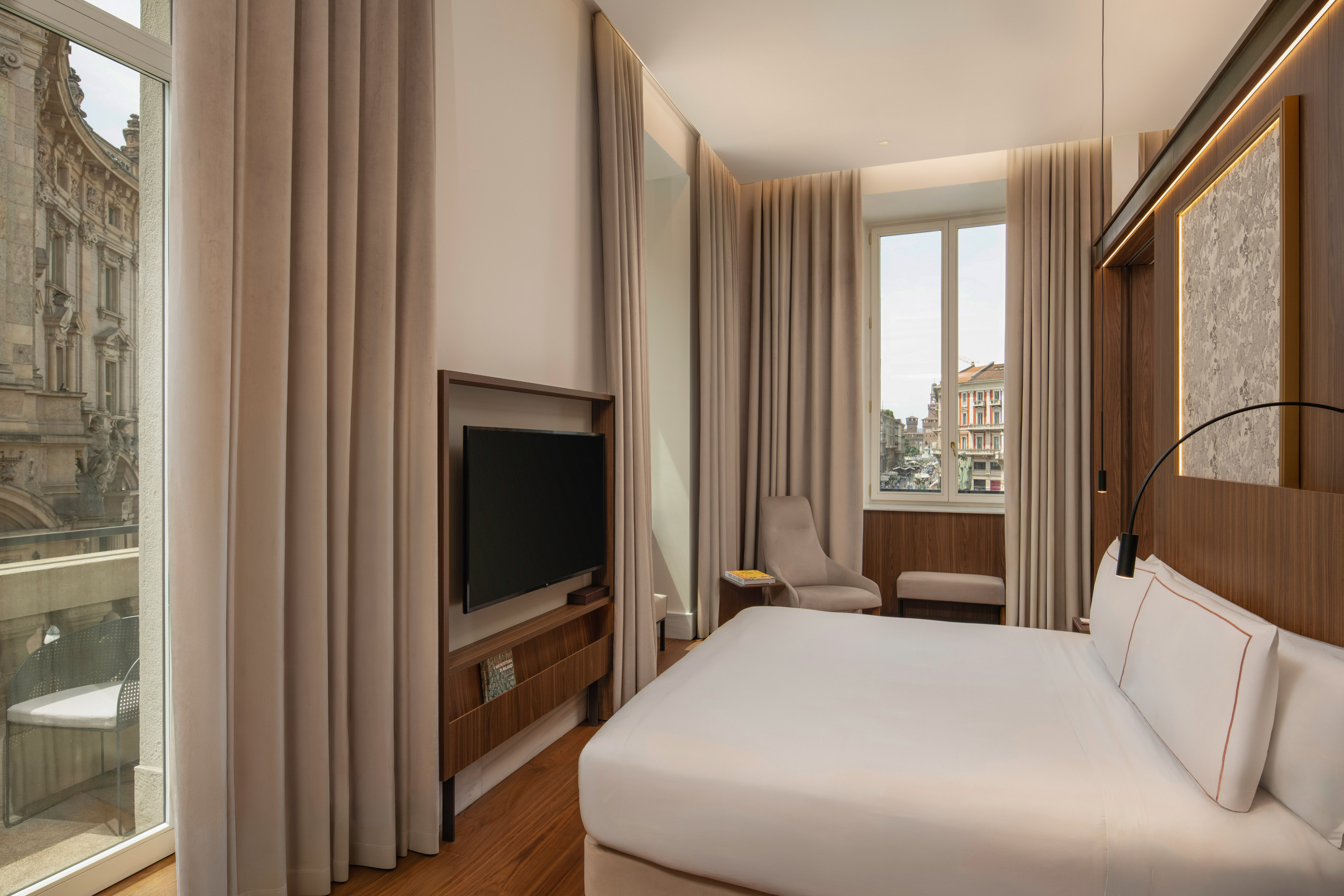 Modern hotel room with king bed, city view, wood floors, and wall-mounted TV.