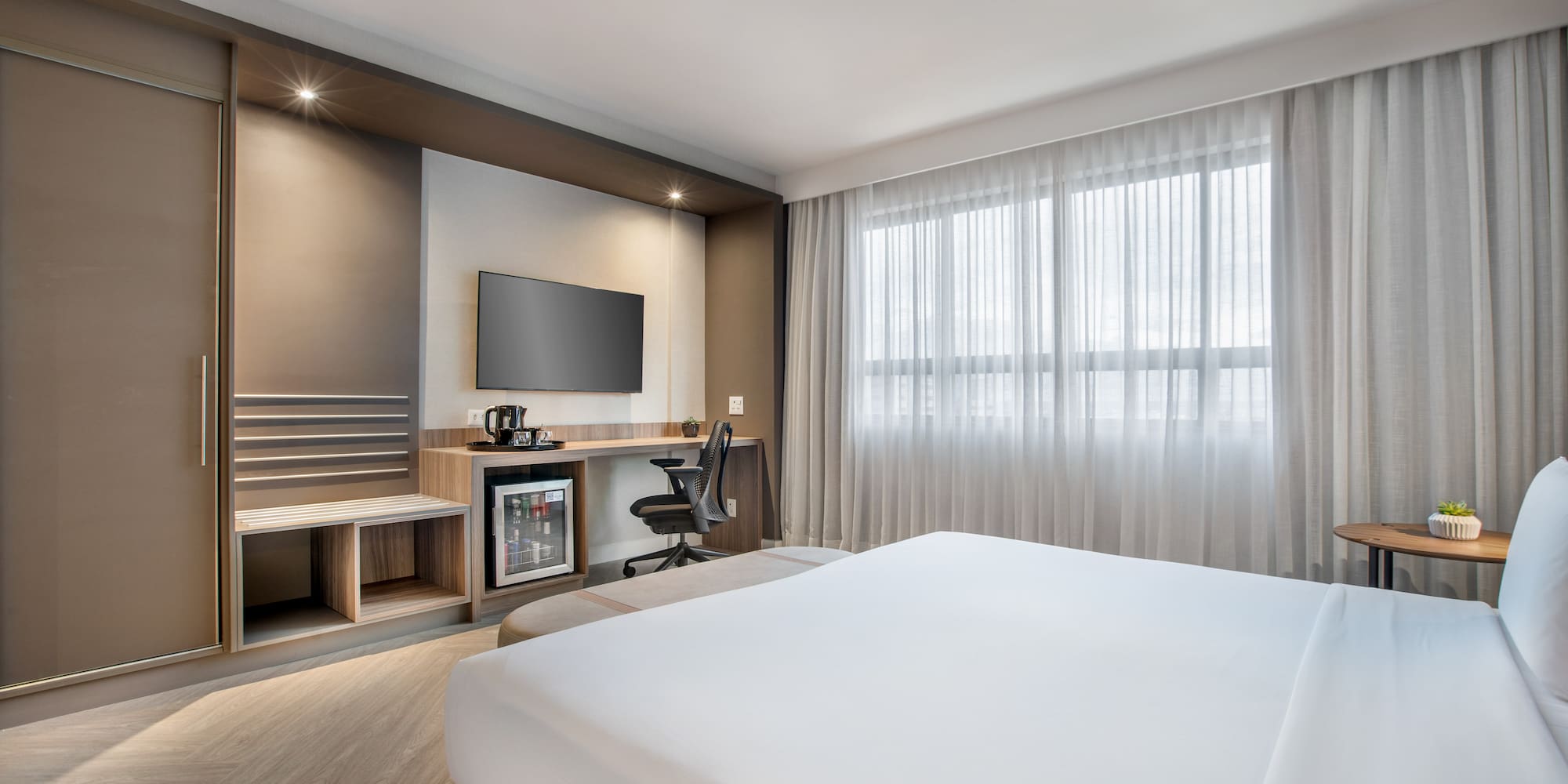 Modern hotel room with bed, desk, TV, minibar, and large window.