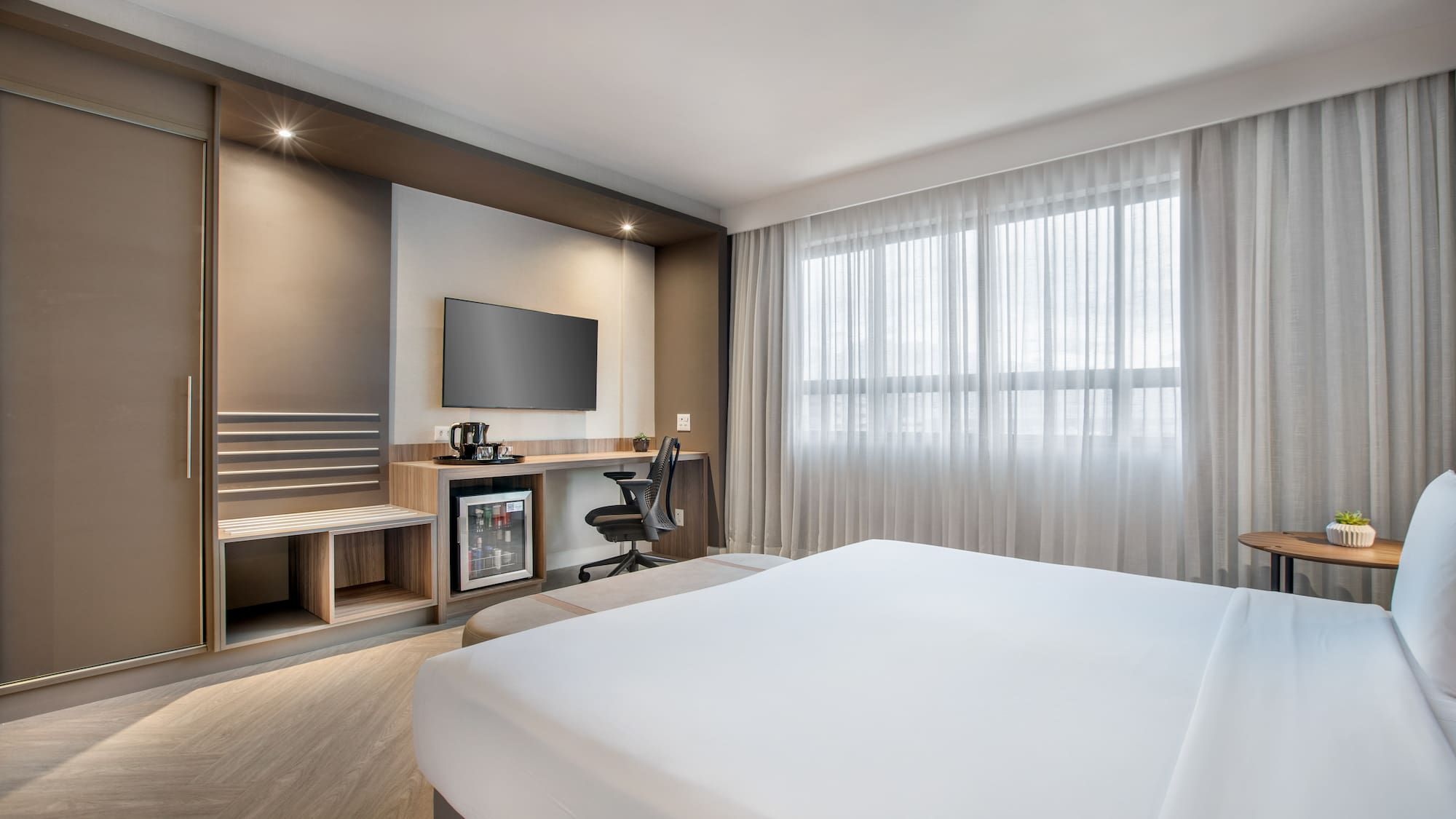 Modern hotel room with bed, desk, TV, minibar, and large window.