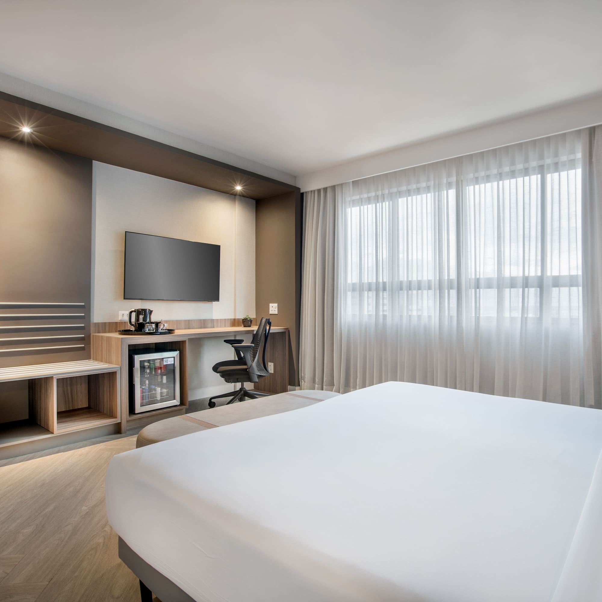 Modern hotel room with bed, desk, TV, minibar, and large window.